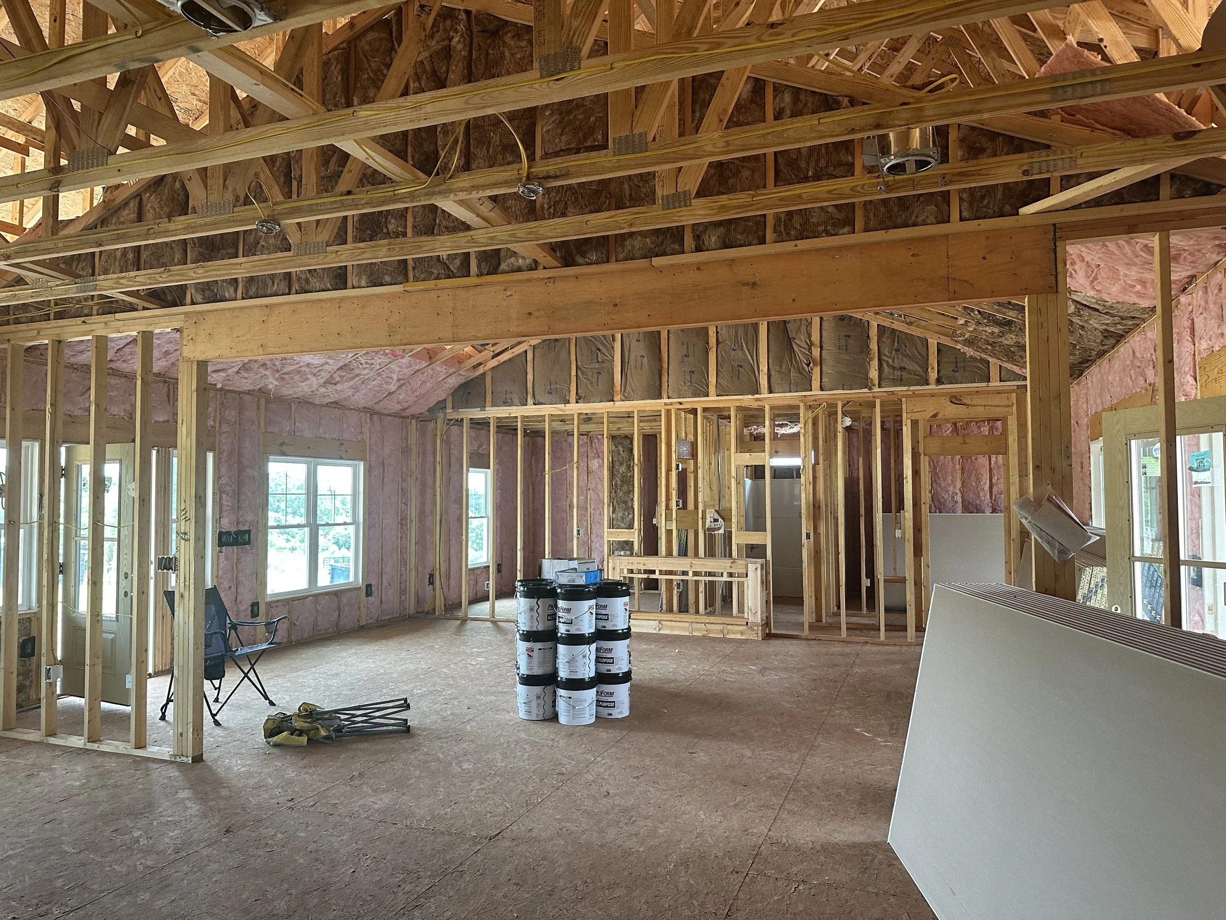 Interior of a house under construction with exposed wooden framing, pink insulation, windows, and construction supplies including buckets and drywall.