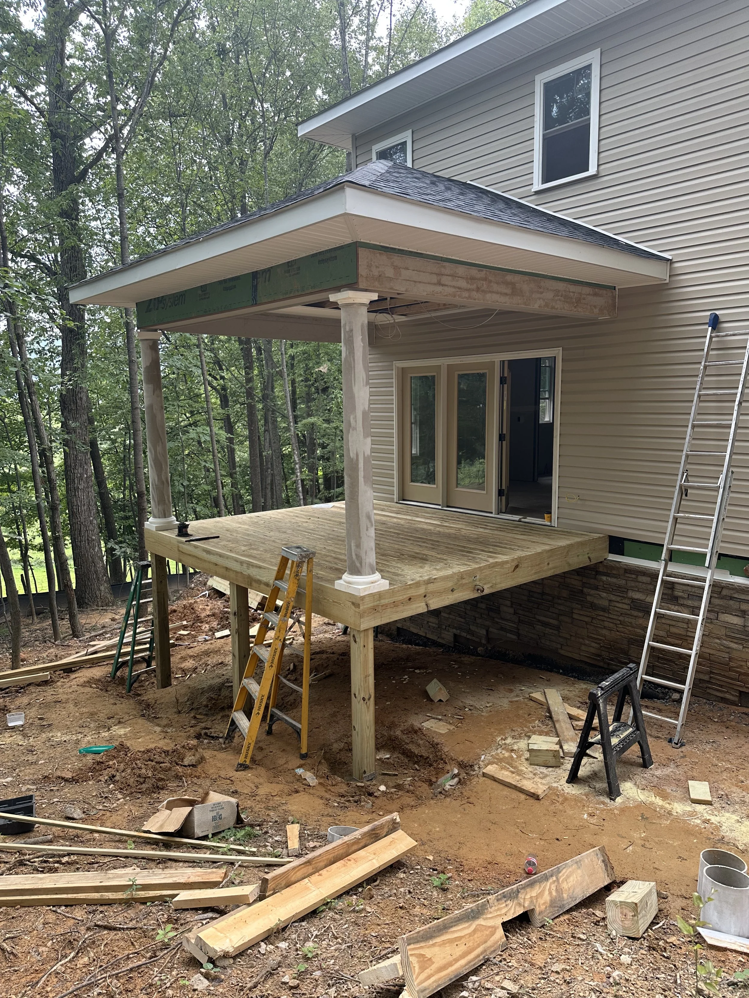 Construction of an outdoor deck with a roof overhang next to a beige house, surrounded by trees, with construction tools and materials on the ground.