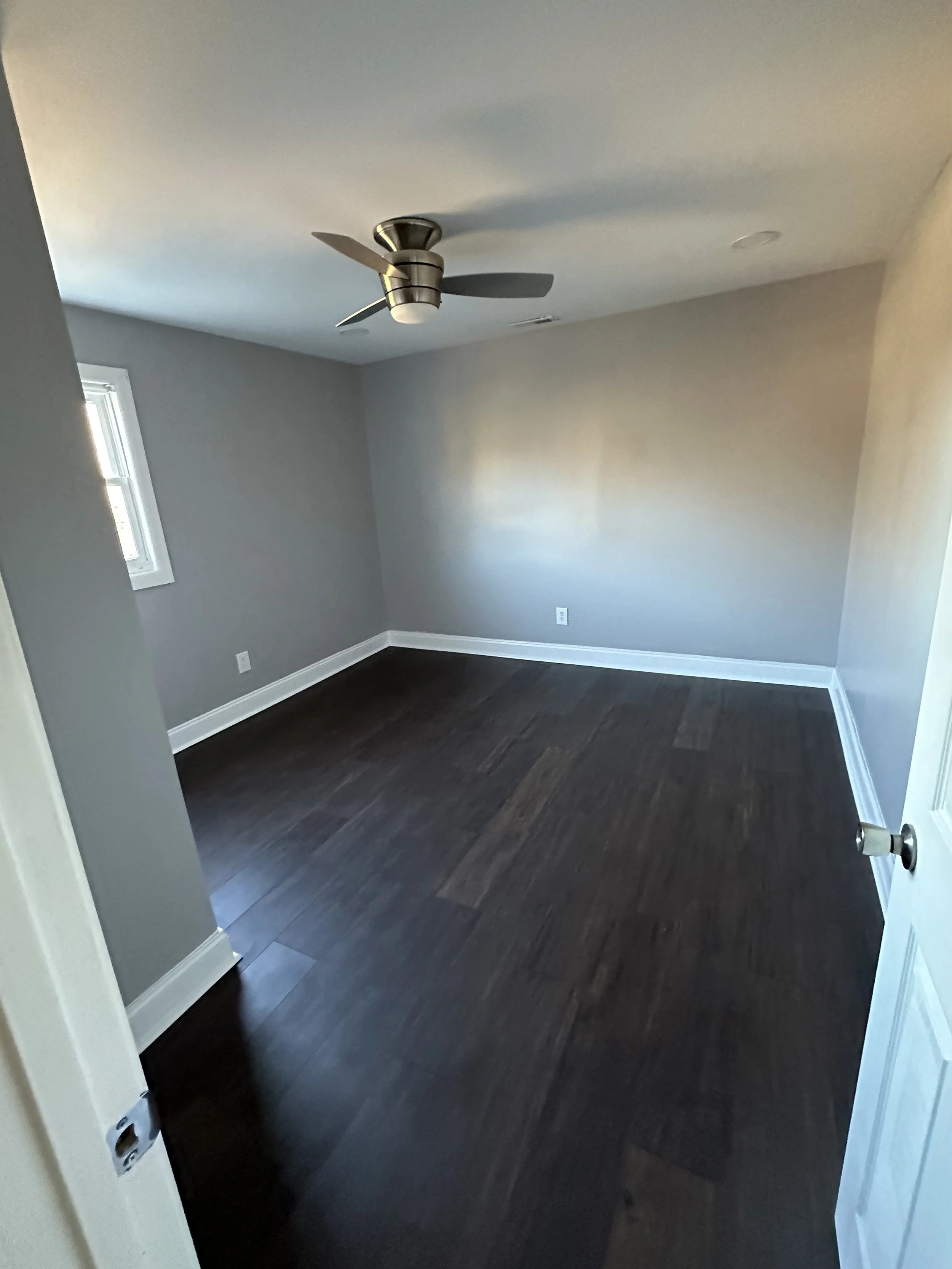 Empty room with dark hardwood floors, light gray walls, a small window, white baseboards, a ceiling fan, and a door slightly open.