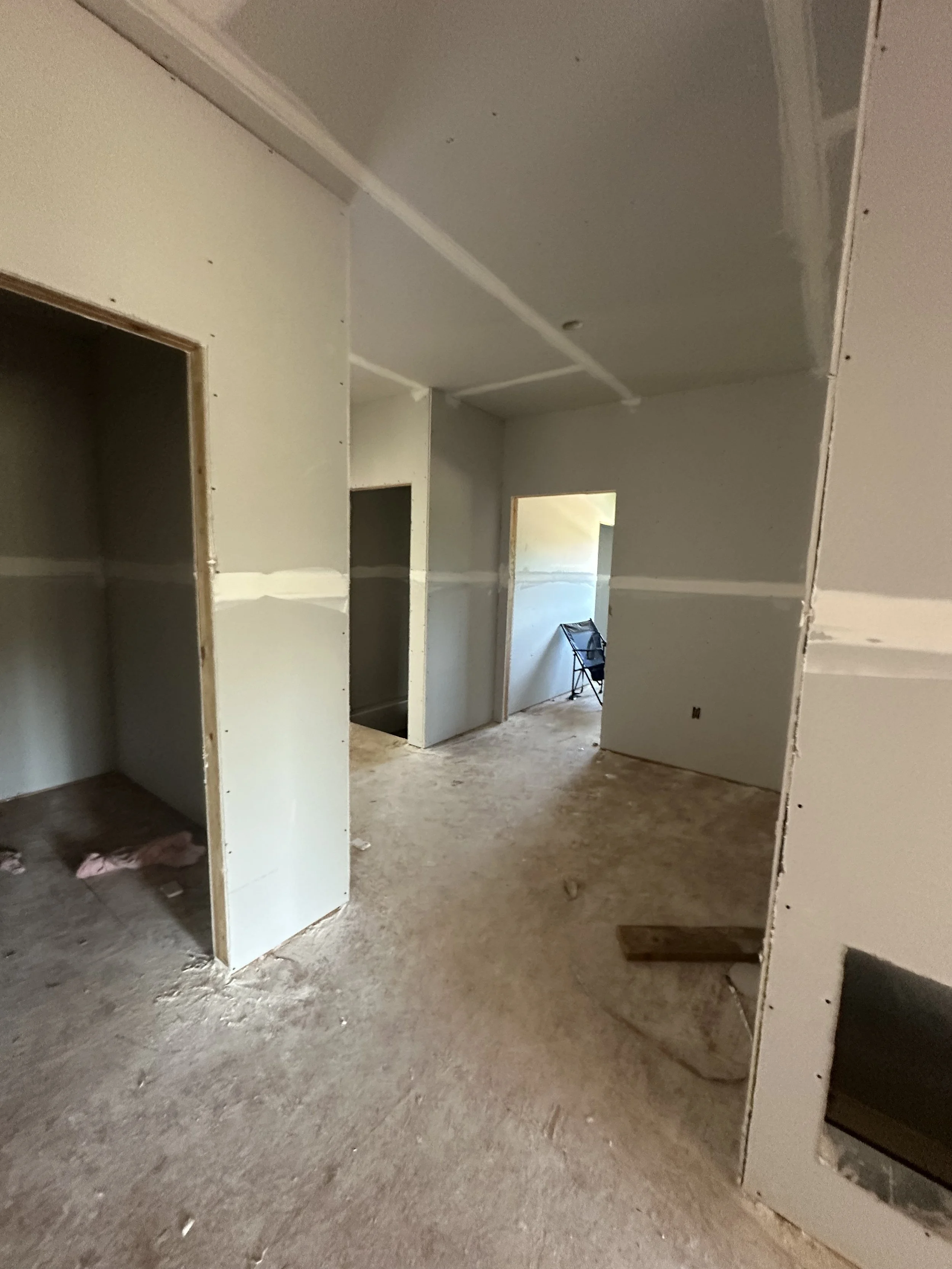 Interior of a room under construction with drywall partially installed, unfinished floors, and an open doorway leading to another area with a chair.