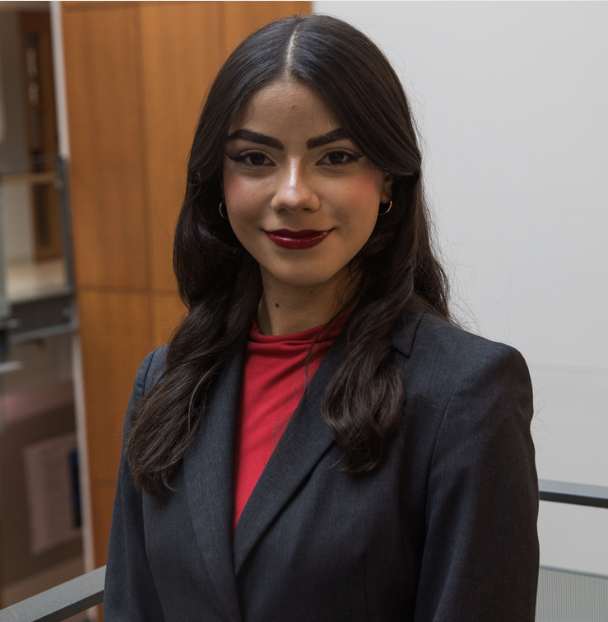 A young woman with dark hair, smiling, wearing a black blazer over a white top, standing indoors with industrial background elements.