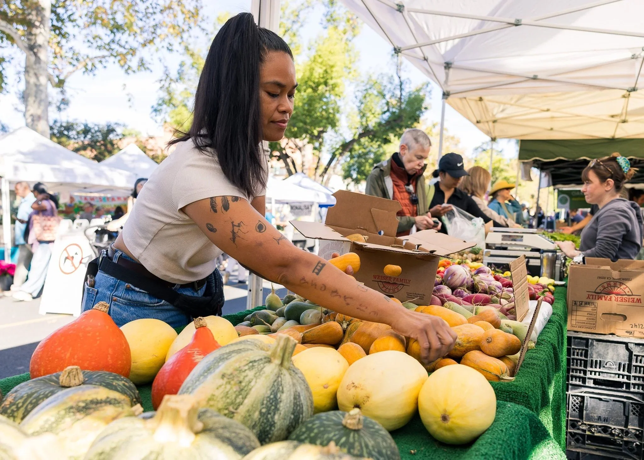 Cat Castaneda with tattoos on her arm shopping for squash and pumpkins at an outdoor farmers market, with other shoppers and vendor tents in the background.