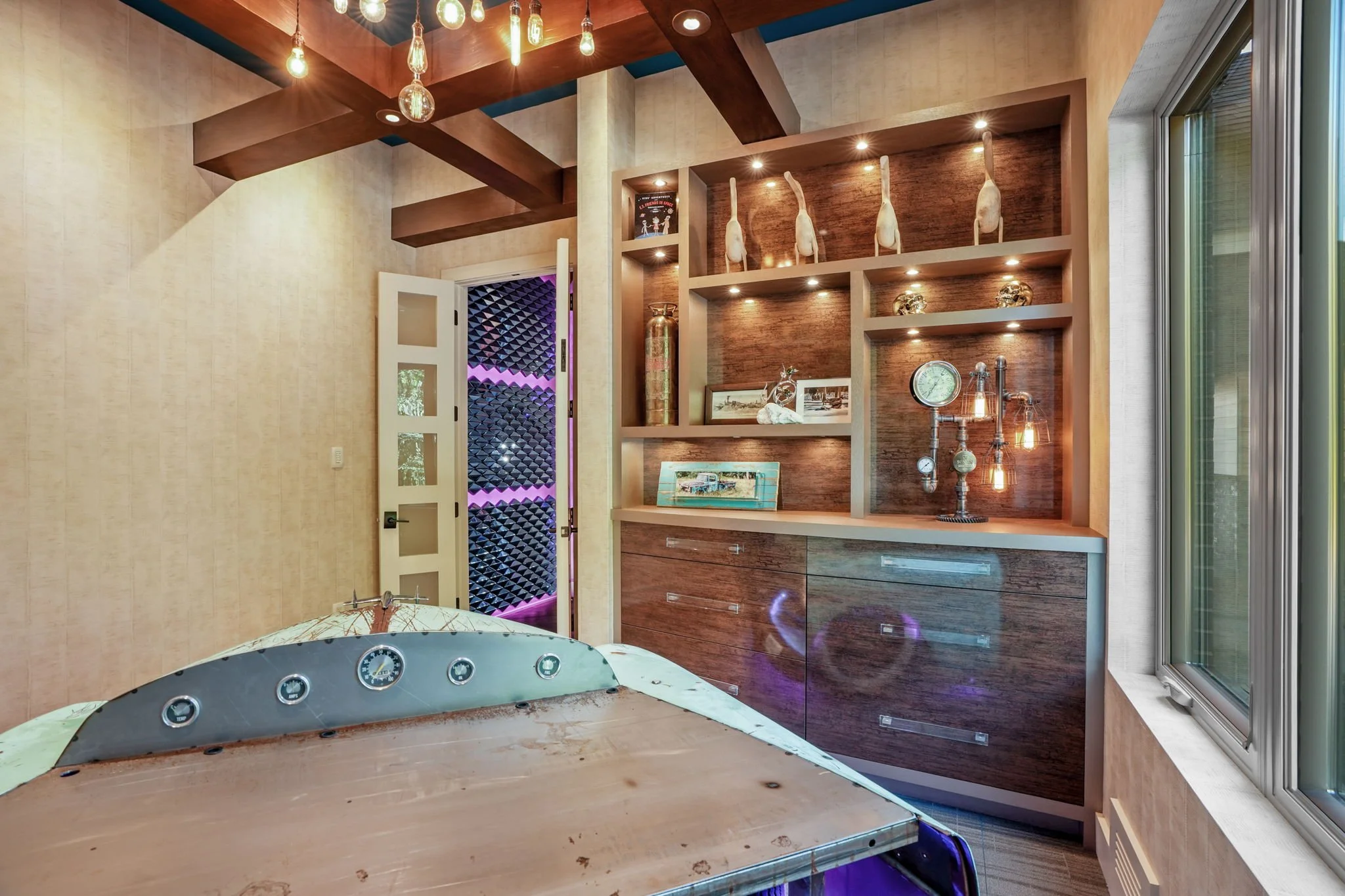 Interior of a room with a bed frame, decorative shelves with art and trophies, modern lighting, and a window showing outdoor greenery.