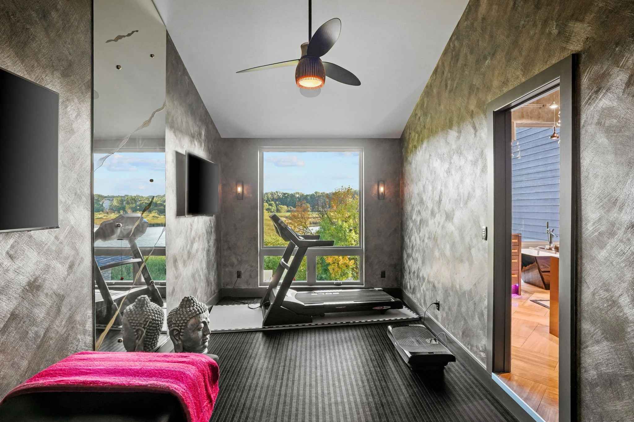 Home gym room with treadmill facing window overlooking green landscape, with two Buddha statues and a table with a pink cloth in the foreground.