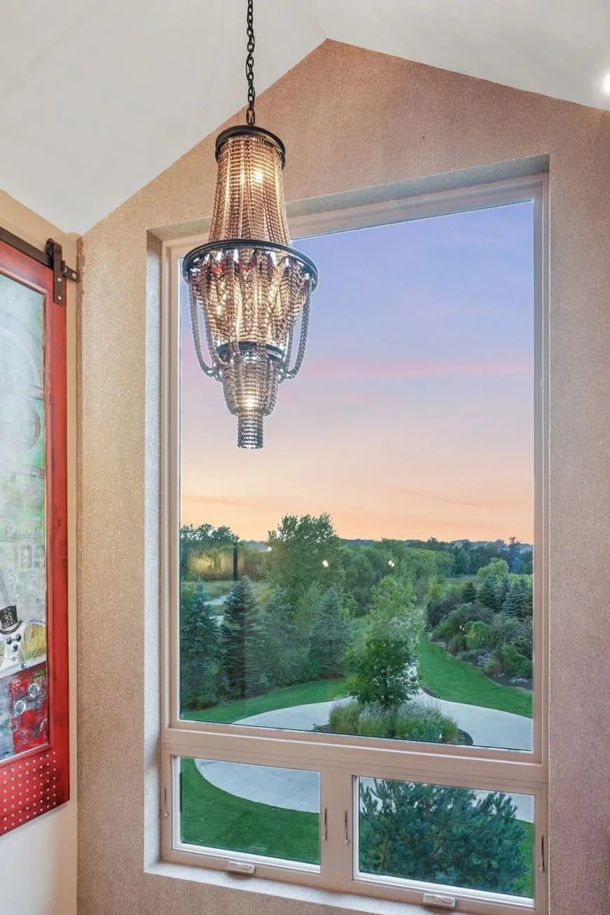 Interior view of a corner with a large window overlooking a park with trees and walking paths at sunset. A decorative chandelier hangs in the upper left corner.