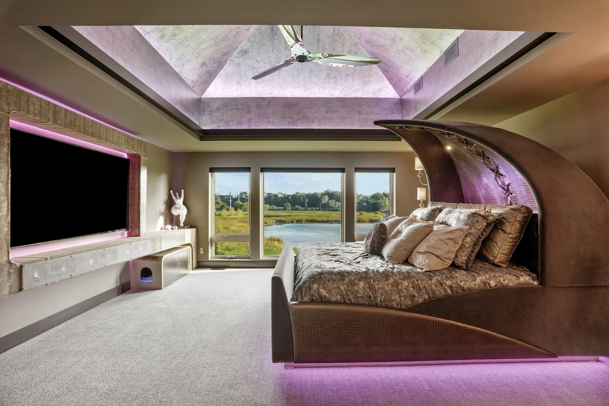 Luxury bedroom with moon-shaped bed, large windows overlooking a lake, flat-screen TV, and a ceiling fan.