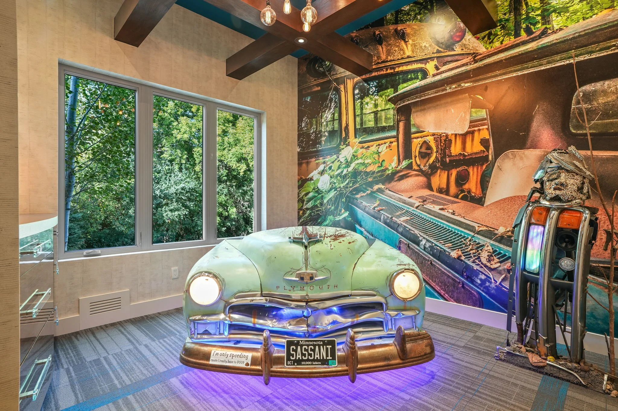 A vintage Pontiac car display with headlights on, Minnesota license plate reading 'SASSANI,' and a humorous sticker that says 'I'm only speeding cause I really have to poop.' The car is in a room with a large window showing green trees outside and a