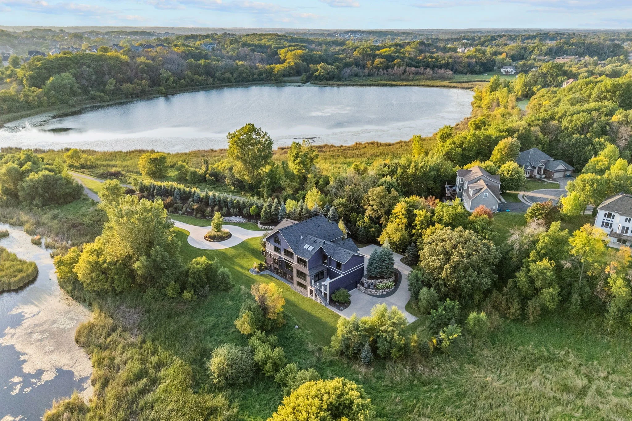 Aerial view of a house surrounded by trees next to a pond, with additional houses and greenery in the background.