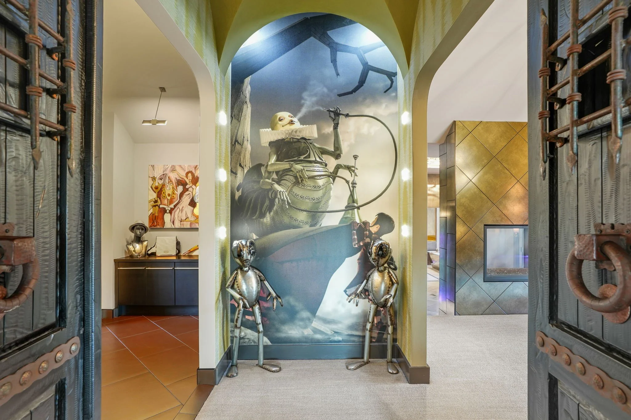 Artistic interior space with a large surreal mural featuring a figure playing a wind instrument, wearing a ruffled collar, surrounded by metallic humanoid sculptures of various sizes, framed by ornate wrought iron gate doors.