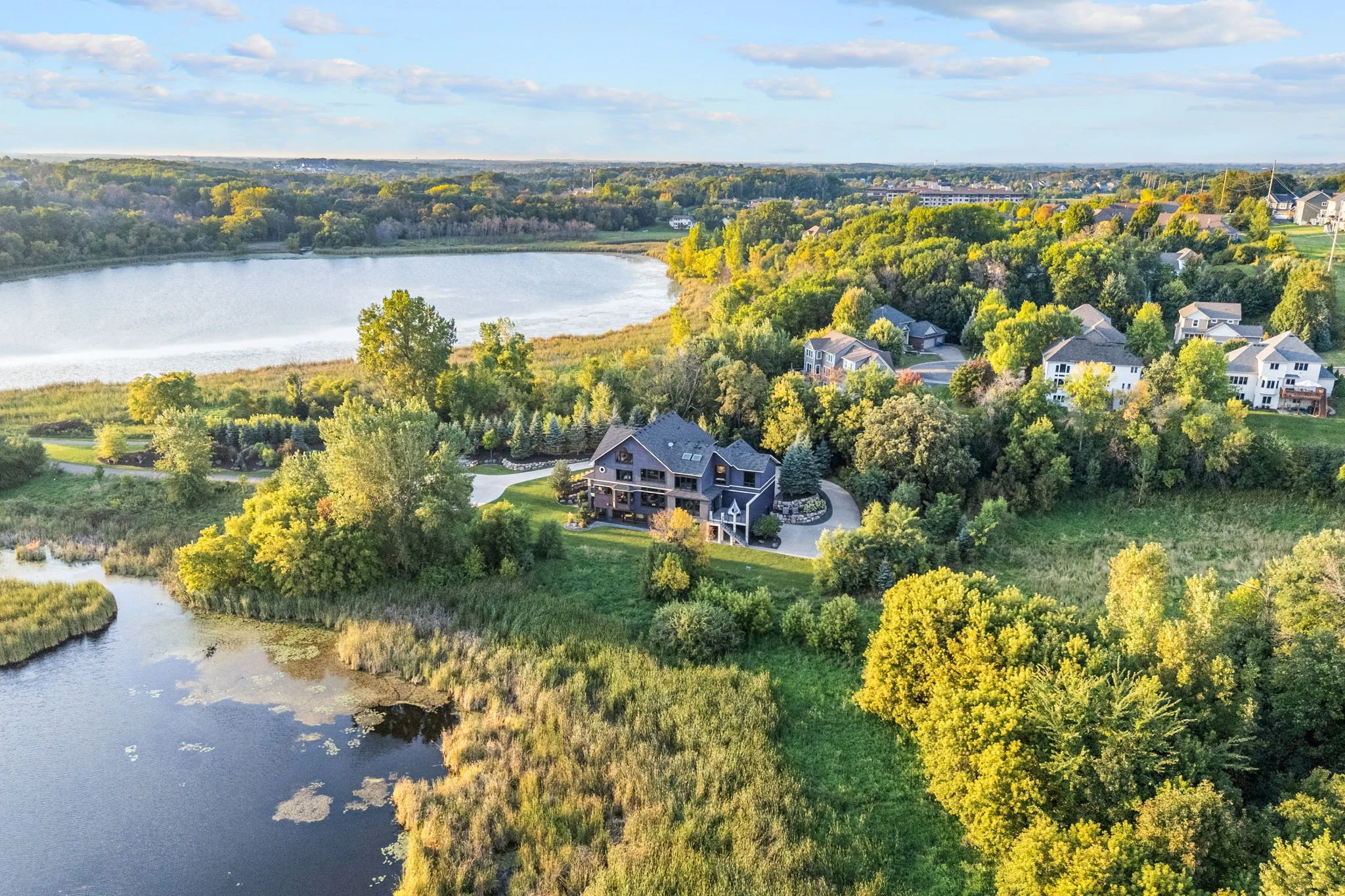 Aerial view of a large house surrounded by trees near a lake with a community of houses in the background.