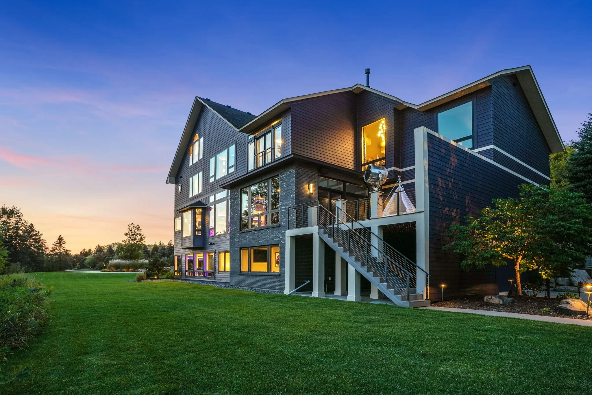 Modern multi-story house with dark siding, large lit windows, and a backyard lawn during dusk, with trees and a colorful sky in the background.