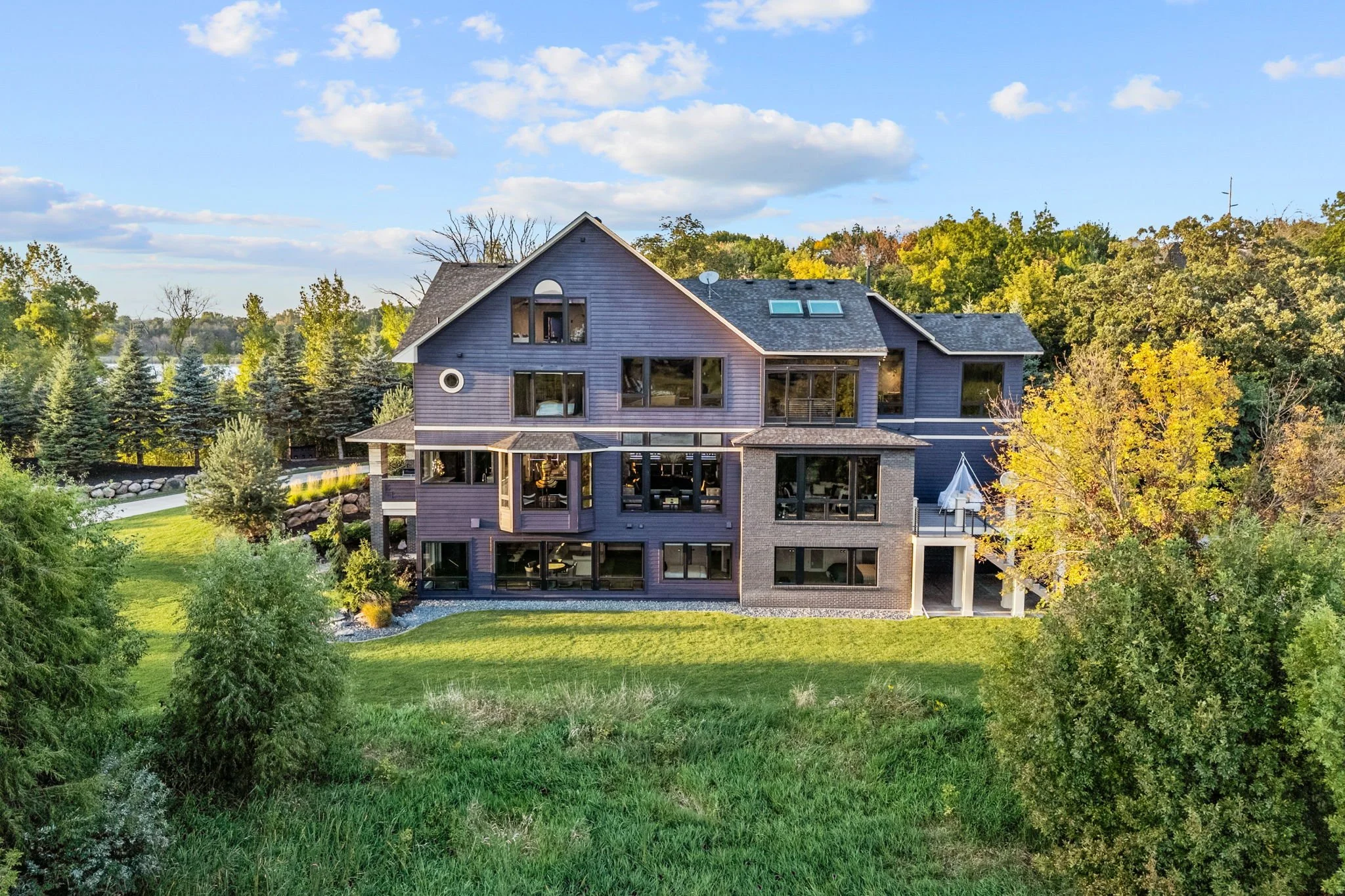 Large modern house with multiple stories, blue exterior, large windows, surrounded by trees and greenery, with a water body in the background, under a partly cloudy sky.