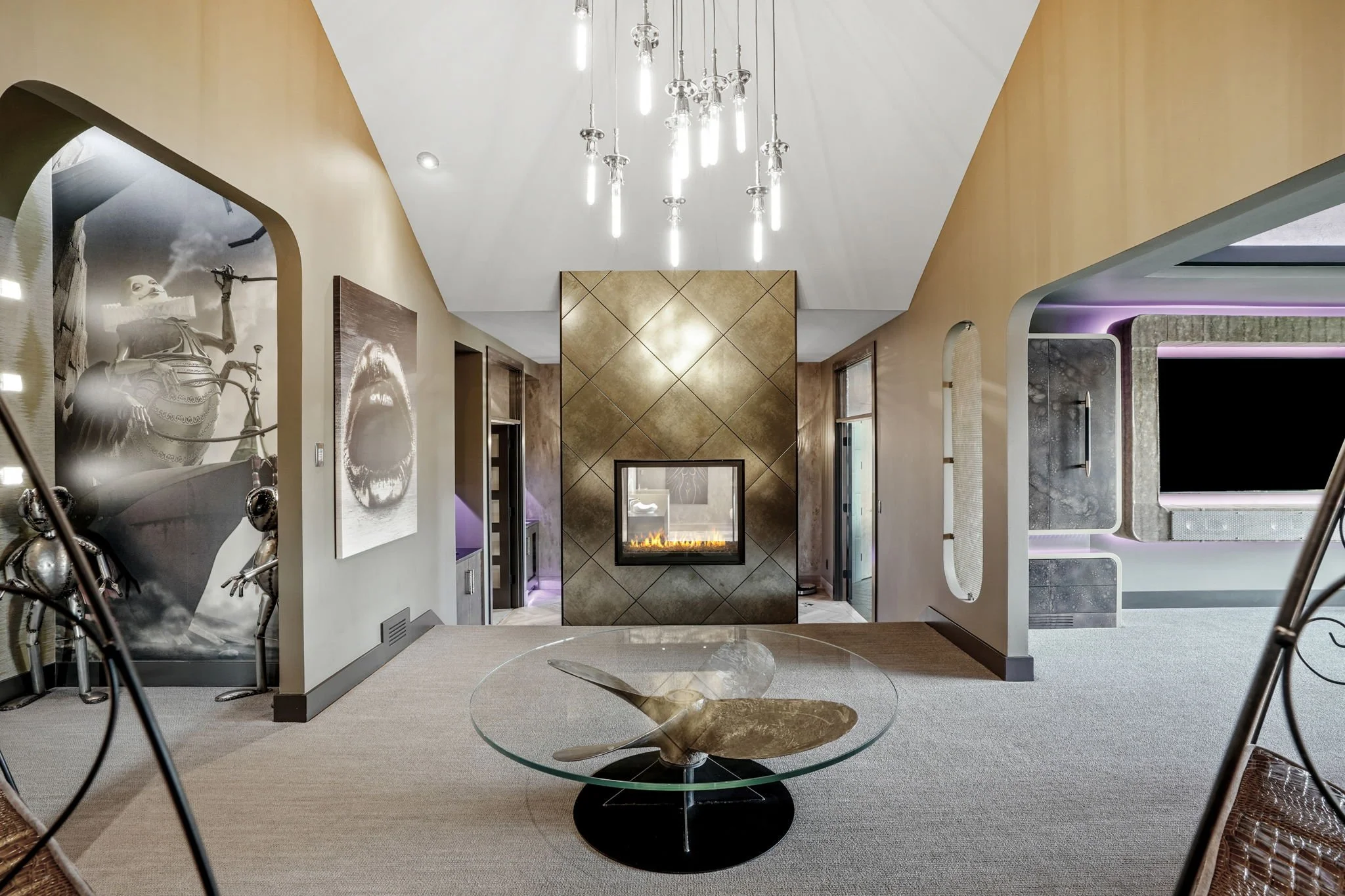 Interior of a modern living room with a central glass coffee table, metallic wall panel with fireplace, chandelier, artwork on walls, and adjacent spaces with purple lighting.