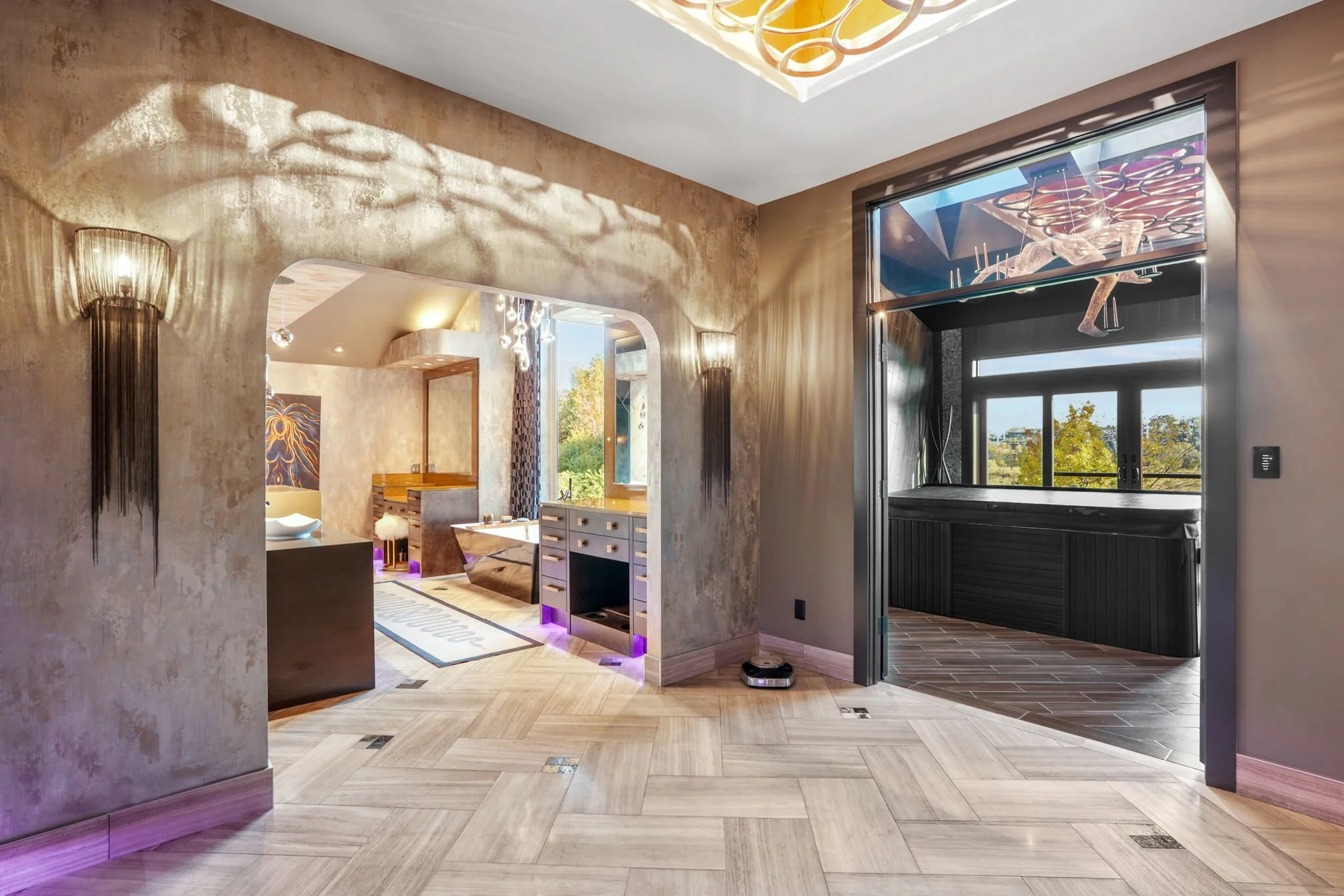 Modern interior living space with textured beige walls, a decorative wall sconce, an arched doorway leading to a room with a bathtub and artwork, and a second room with large windows, a hot tub, and a geometric ceiling installation. Light wood floori