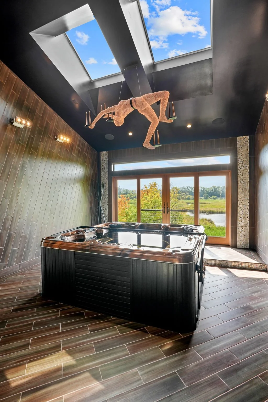 A room featuring a hot tub with a panoramic view through large glass doors, a ceiling skylight, and a sculpture of an acrobat hanging from the ceiling.