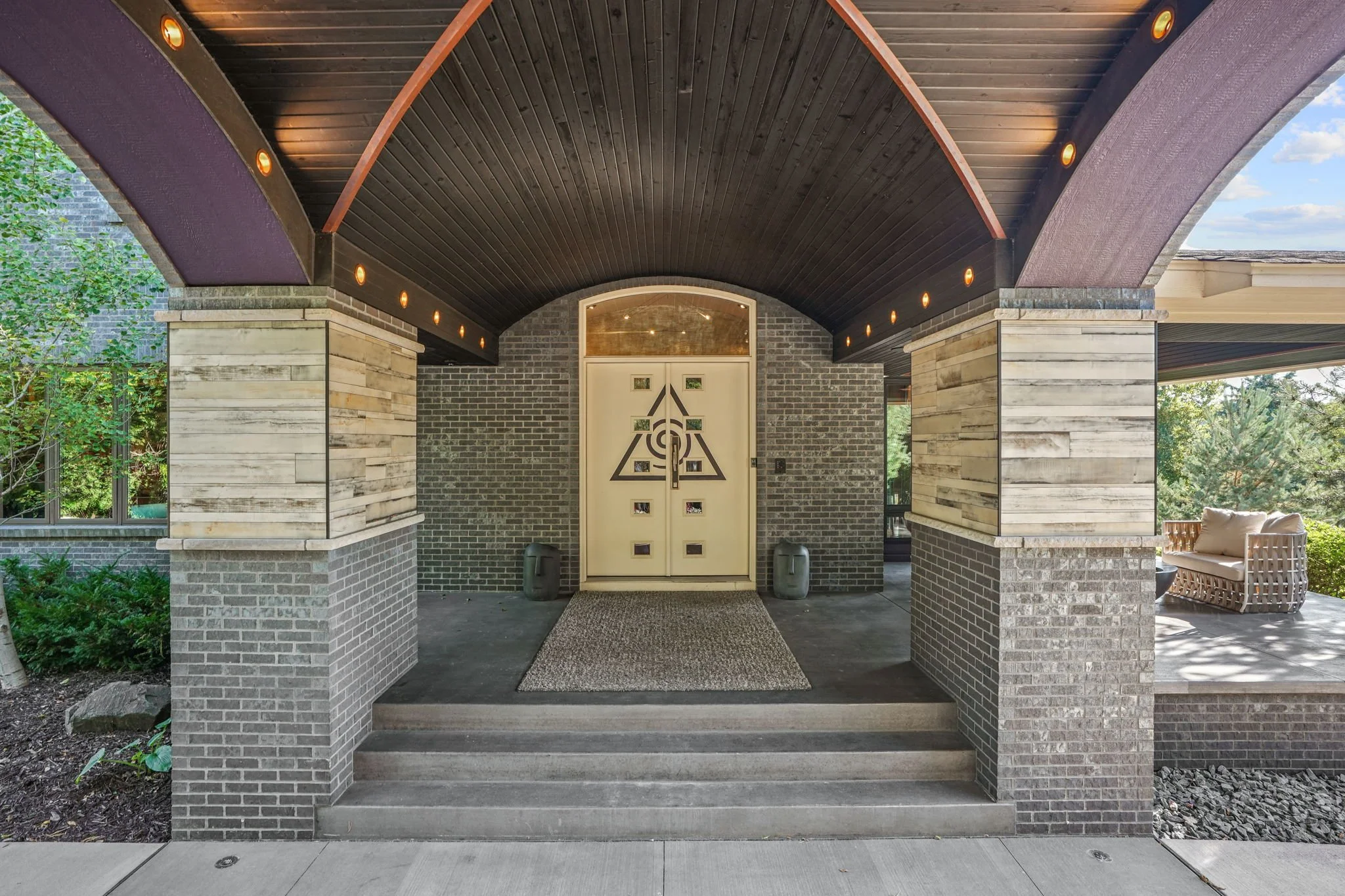 Front porch of a modern house with a dark arched ceiling, gray brick walls, and large wooden columns. There is a beige door with geometric and spiral design, flanked by two large planters, and a small doormat on the concrete steps. To the right, ther