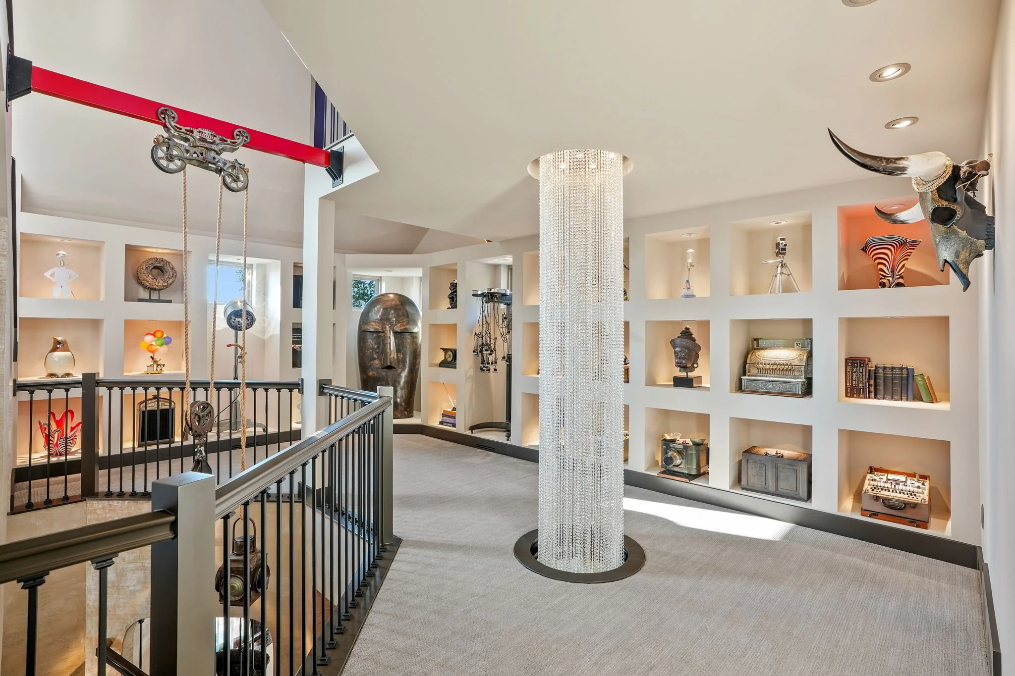 Modern interior of a home with built-in wall shelves filled with decorative objects, a large hanging chandelier, and art pieces including a large face sculpture and animal horns.