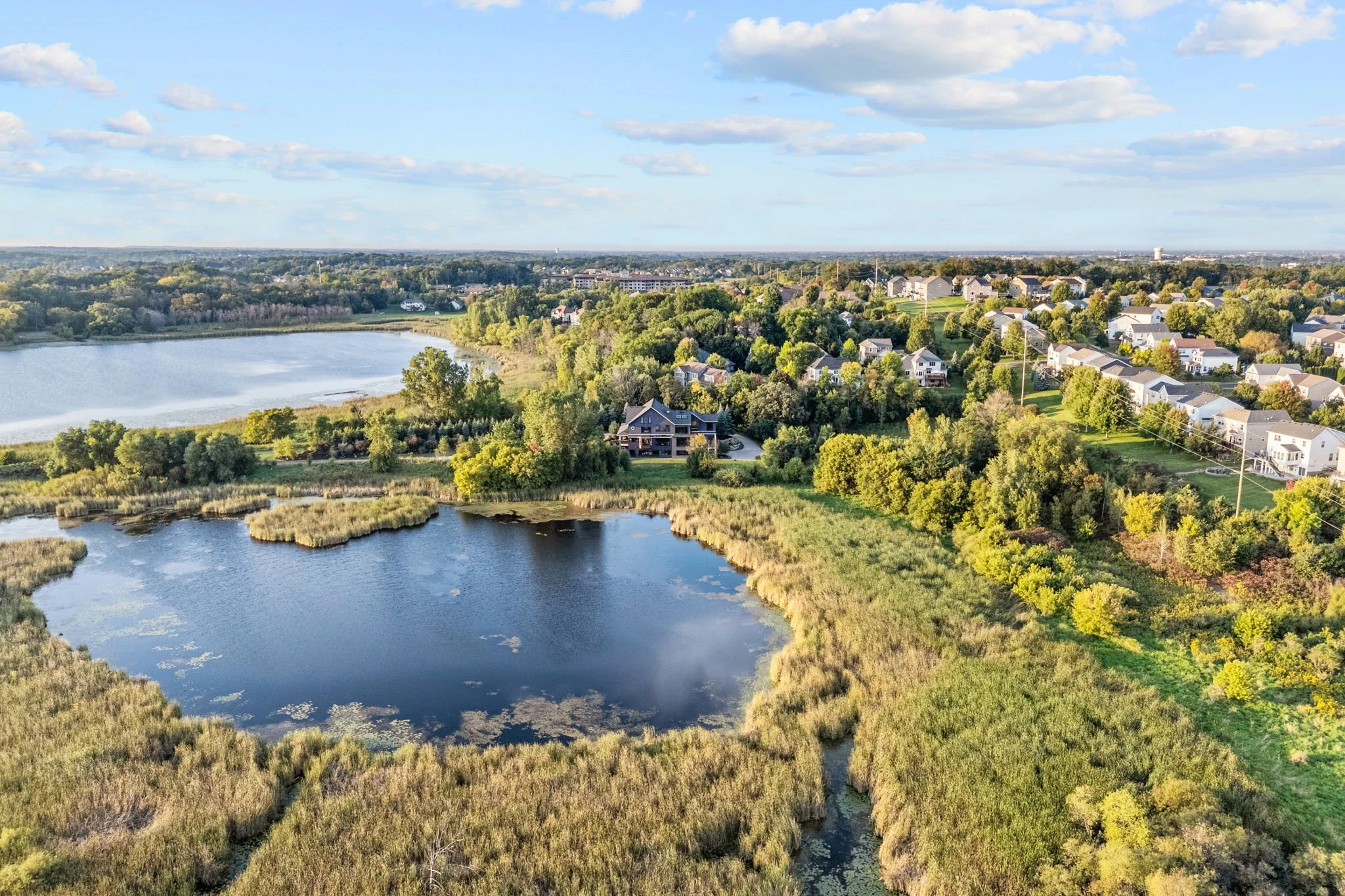 Aerial view of a suburban neighborhood next to a lake with lush green trees and houses in the background under a partly cloudy sky.