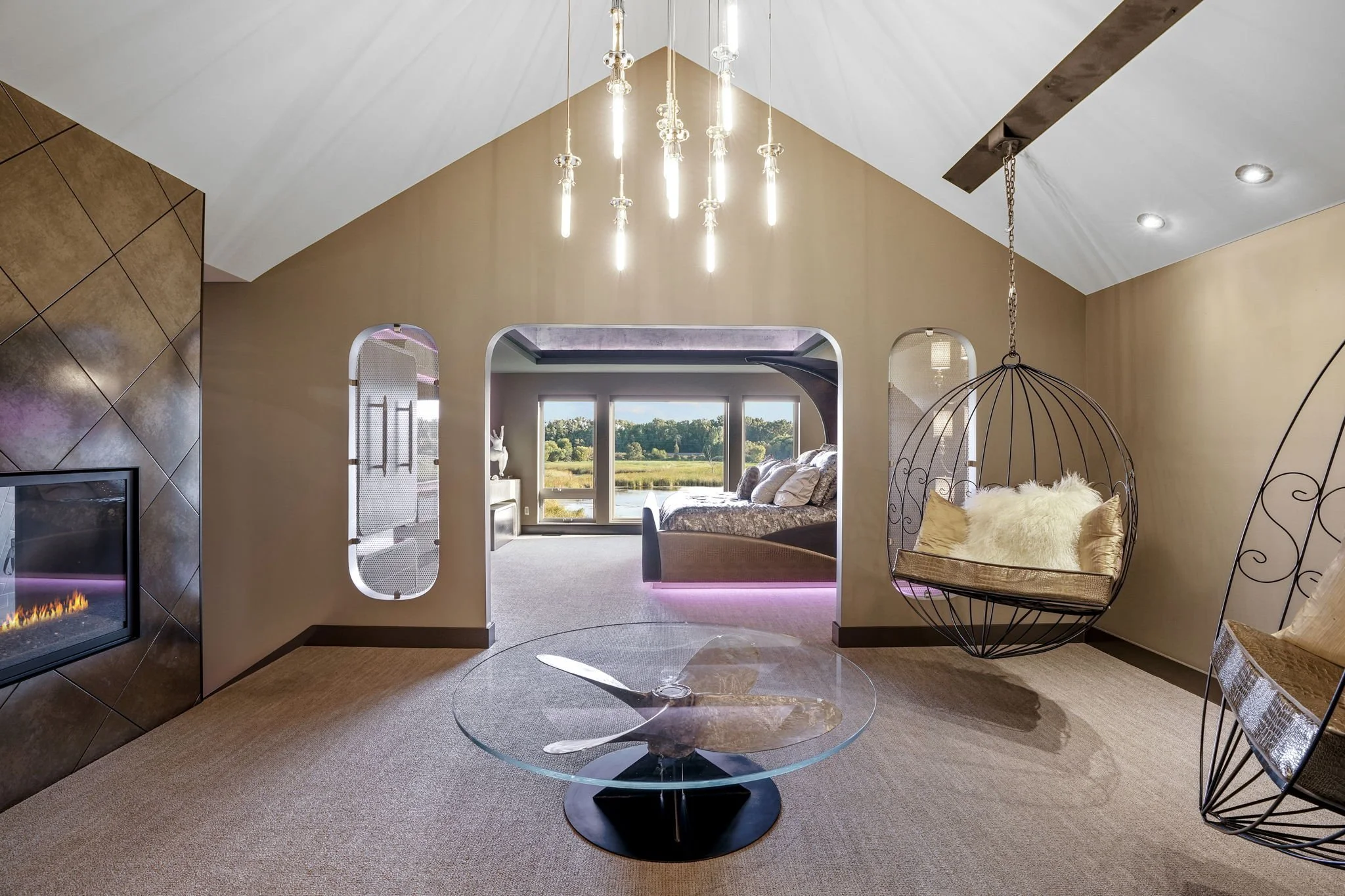 Modern living room with fireplace, hanging spherical chair, glass coffee table, and bed with large windows showing outdoor landscape.