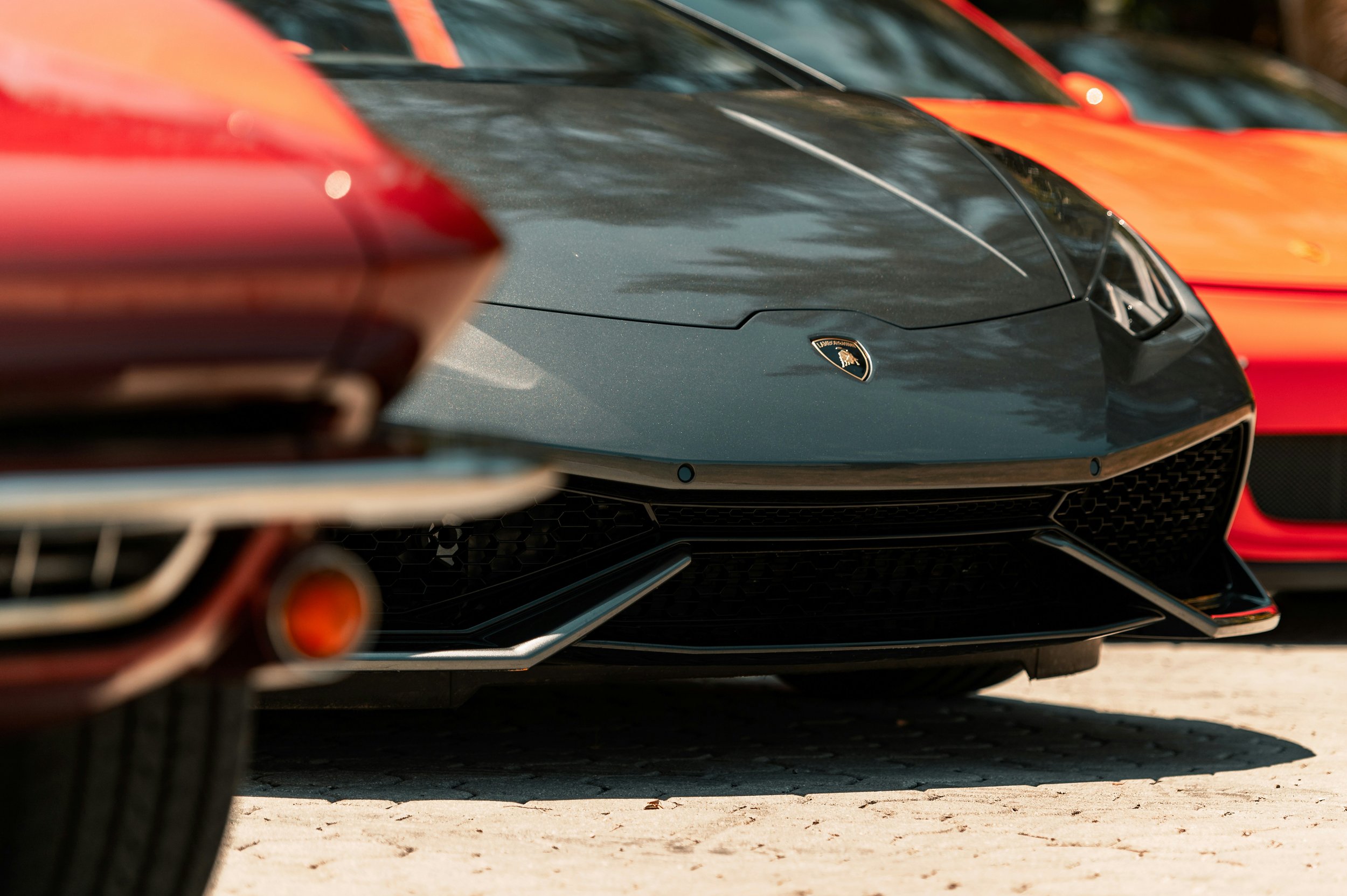 Close-up of the front of a dark gray Lamborghini sports car parked outdoors, with part of an orange car visible on the right and blurry red car in the foreground.