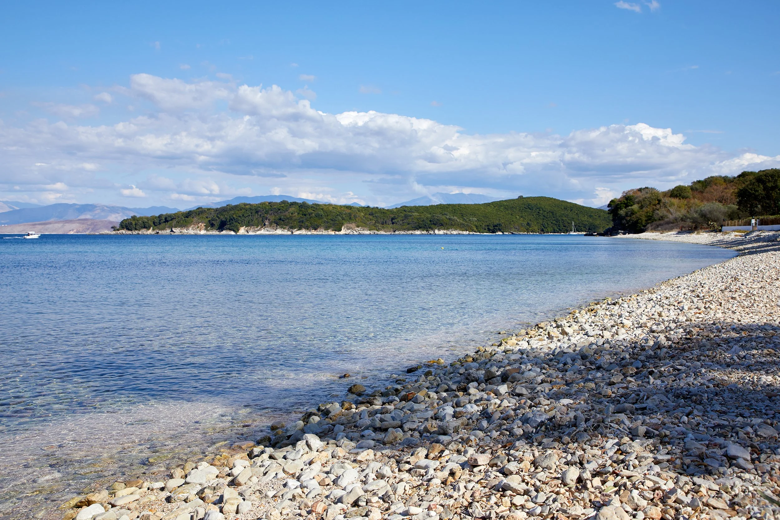 A peaceful beach with clear blue water and a pebble shoreline. In the background, there are green, tree-covered hills under a partly cloudy sky.