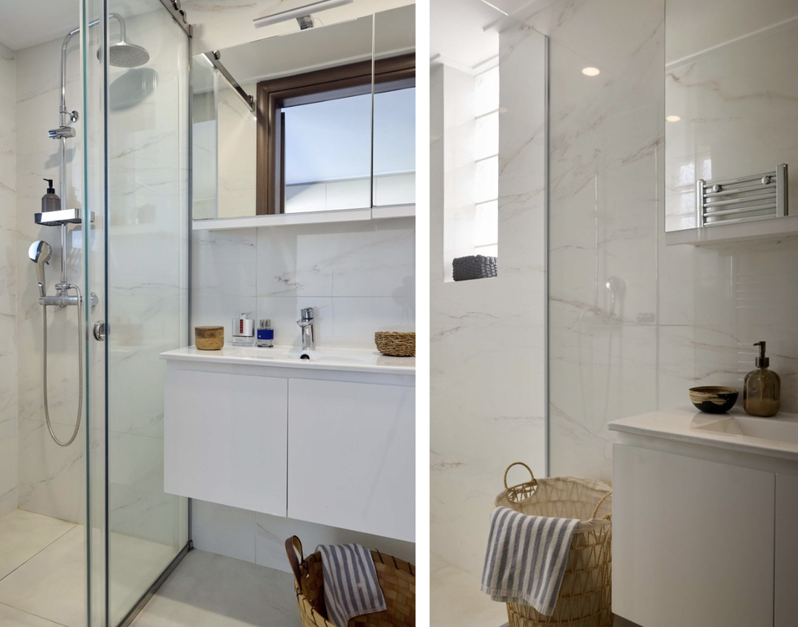 Modern bathroom with a glass-enclosed shower, white vanity, marble walls, towel basket, and a small window.