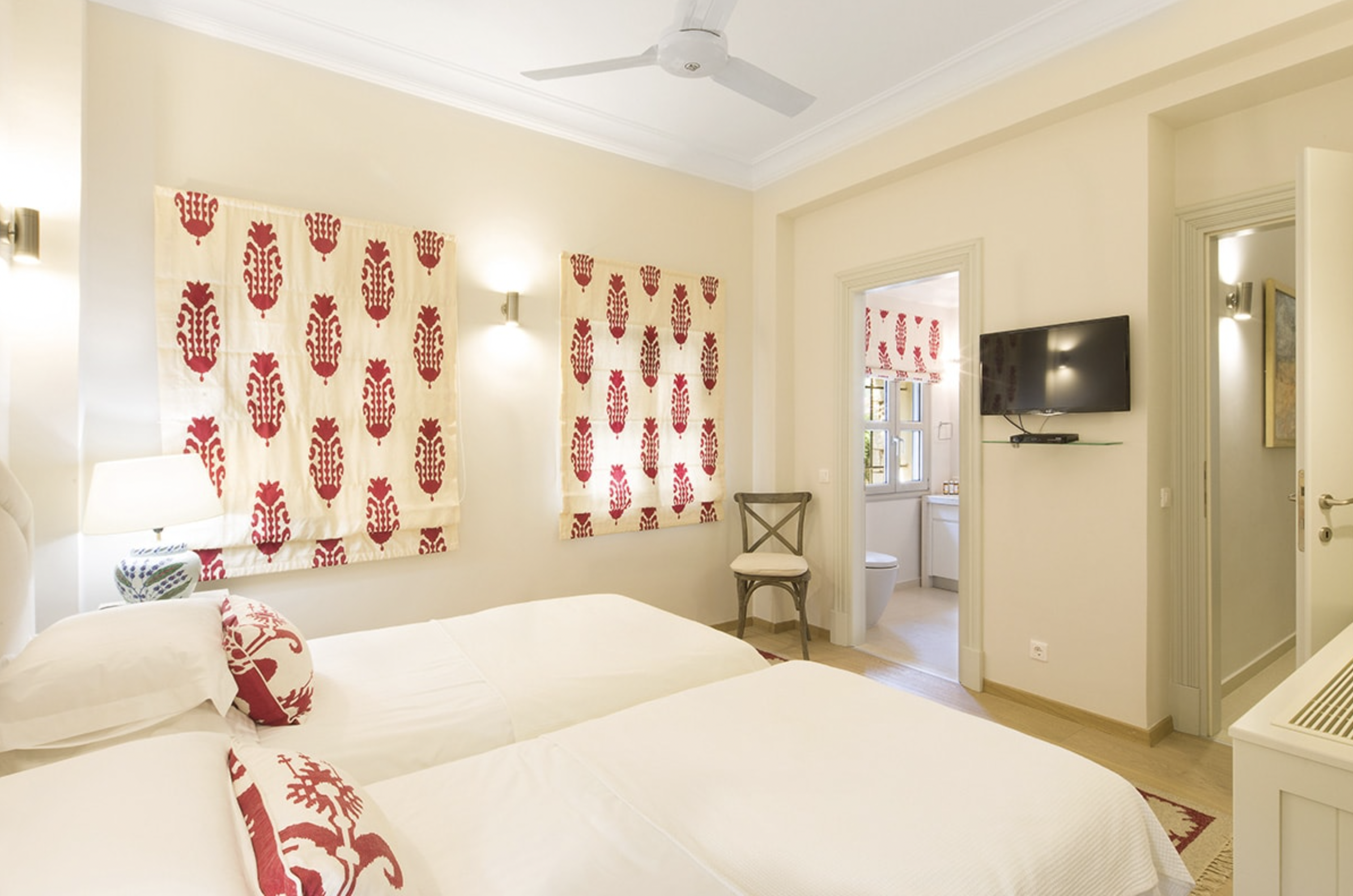 Hotel room with two beds, red and white patterned curtains, wall-mounted TV, and an adjoining bathroom with a window.