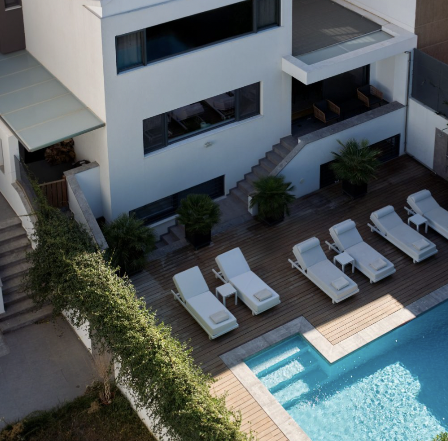 Aerial view of a modern hotel pool area featuring white lounge chairs, small side tables, potted palm trees, a wooden deck, and a swimming pool with steps.