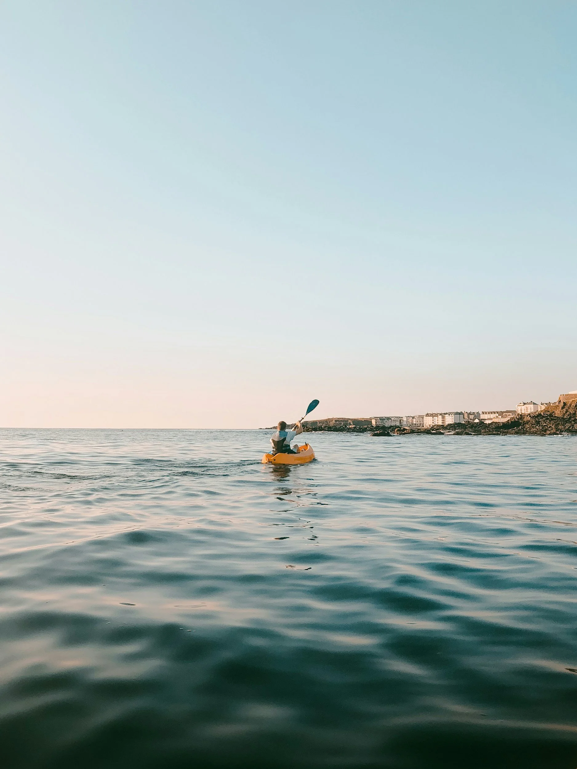 Person in a yellow kayak paddling on calm water near a coastline with buildings in the distance during sunset or sunrise.