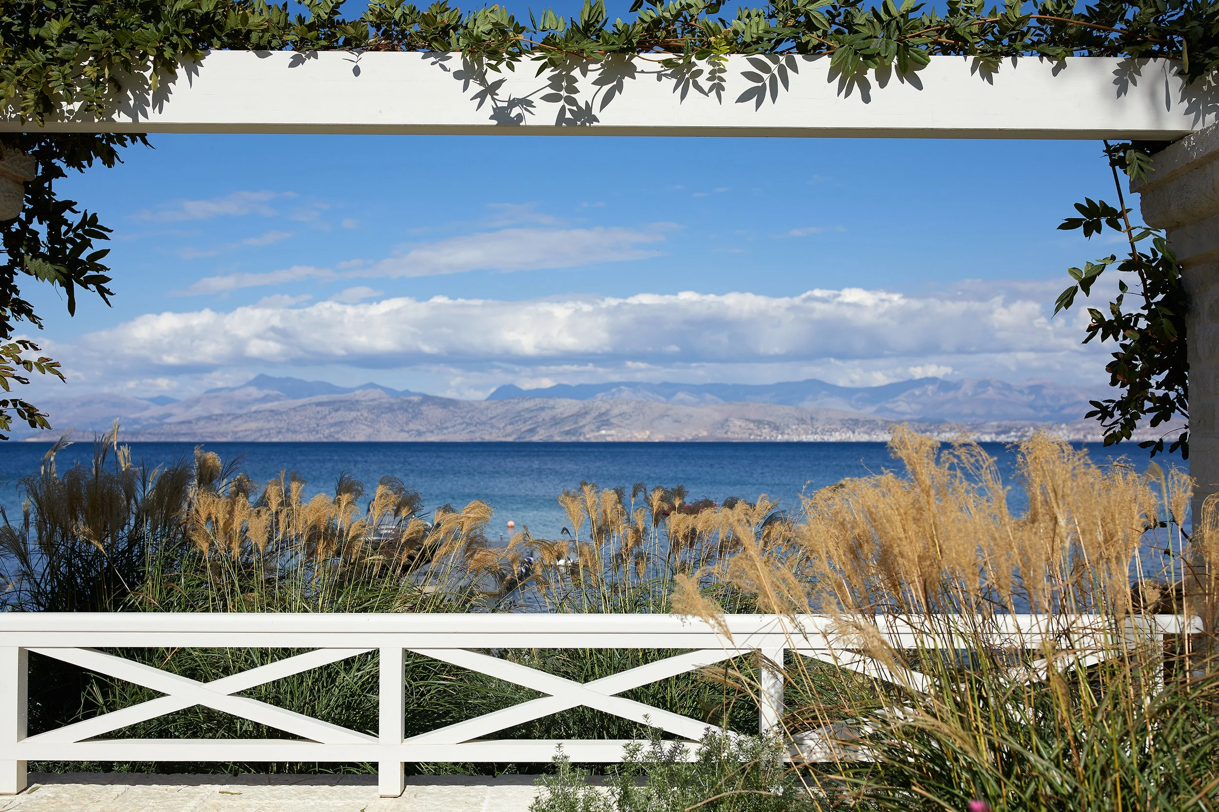 View of a lake with mountains in the distance, framed by a white fence and greenery in the foreground, under a partly cloudy sky.