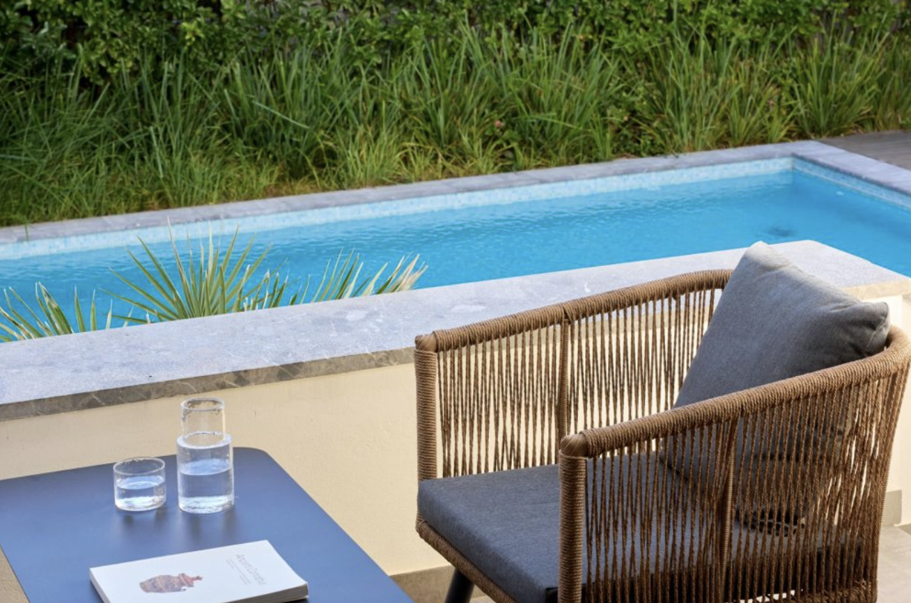 Outdoor patio with a wicker chair, a gray pillow, a table with a book and glasses of water, overlooking a private pool and green plants.