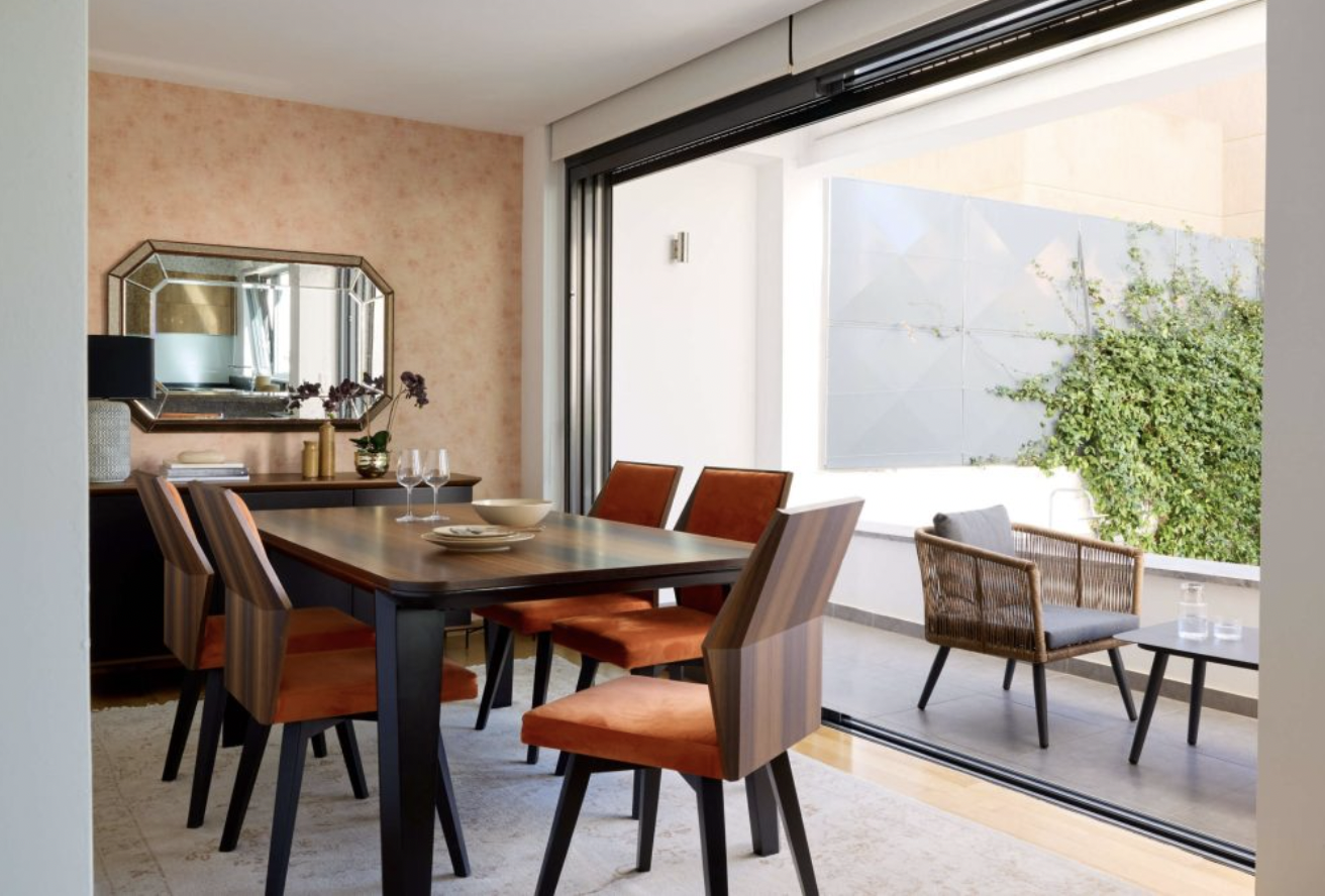 Dining room with a wooden table, six orange cushioned chairs, a sideboard with decor, a large mirror, and an outdoor patio with a wicker chair and small table