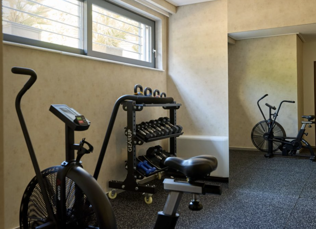 Indoor gym room with exercise bike, dumbbells, and weighted kettlebells, illuminated by natural light from a window.
