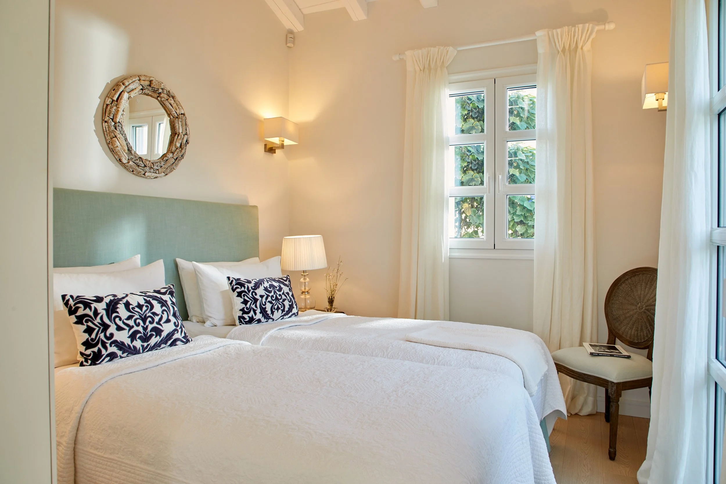 A cozy bedroom with two beds, a nightstand with a lamp, a round mirror on the wall, a wooden chair with booklets, and large windows with white curtains letting in natural light.