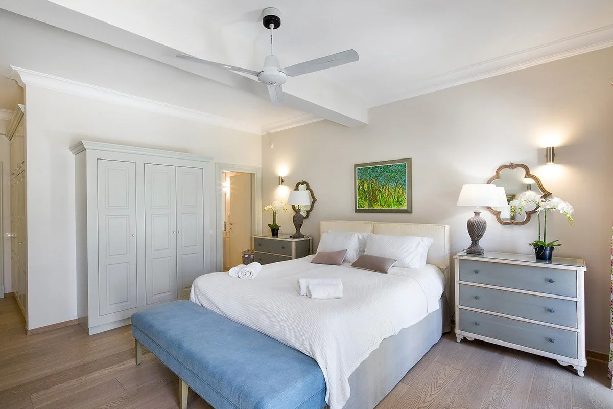 A neatly made bed with white bedding and two brown pillows, flanked by two matching nightstands with lamps and decorative plants, in a bright, cozy bedroom with white walls, a ceiling fan, and a painting above the bed.