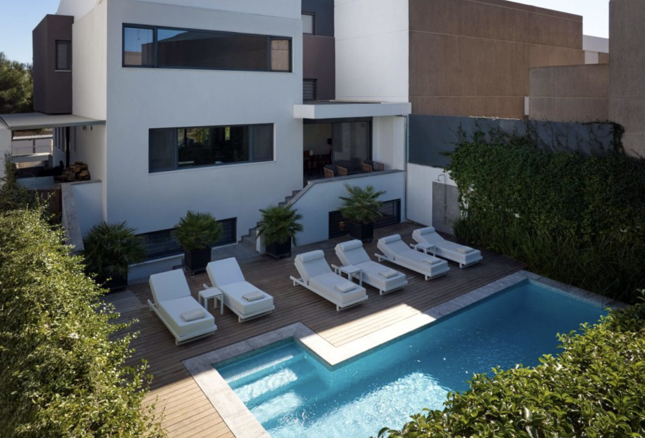 Modern house with a pool and outdoor lounge area featuring white lounge chairs, potted plants, and a backyard enclosed by greenery and tall walls.