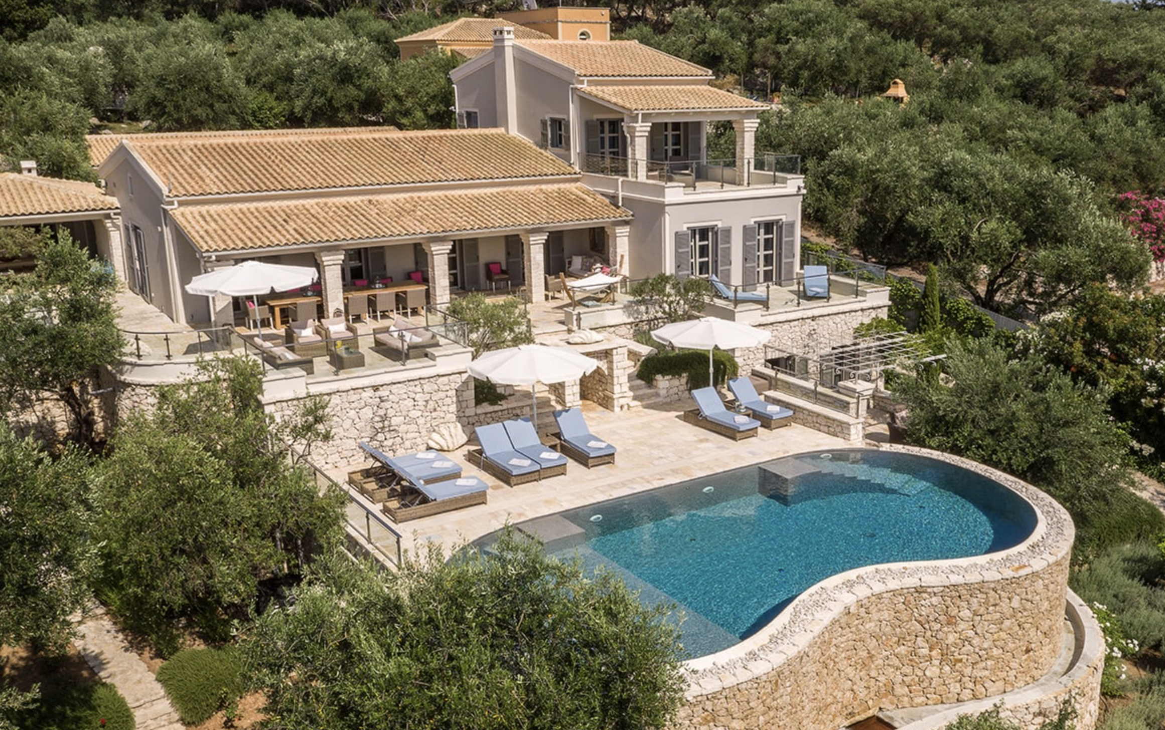 Luxury house with multiple outdoor patios, umbrellas, lounge chairs, and a curved swimming pool surrounded by greenery and trees