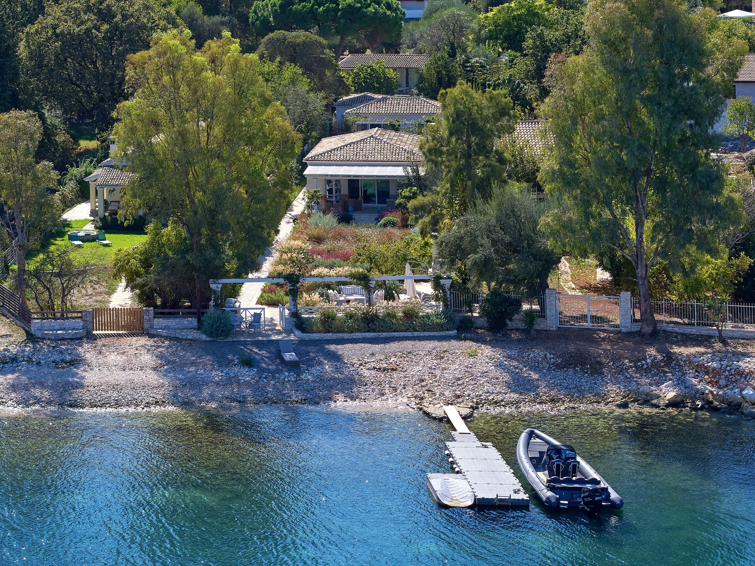 A lakeside house with a garden, trees, and a patio. There is a dock extending into the water with a boat attached.