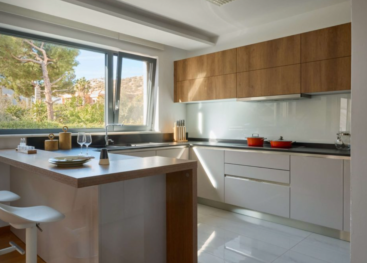 Modern kitchen with white cabinets, wooden upper cabinets, black countertop, large window showing outdoors, red pots on stove, and dining area with plates and wine glasses.
