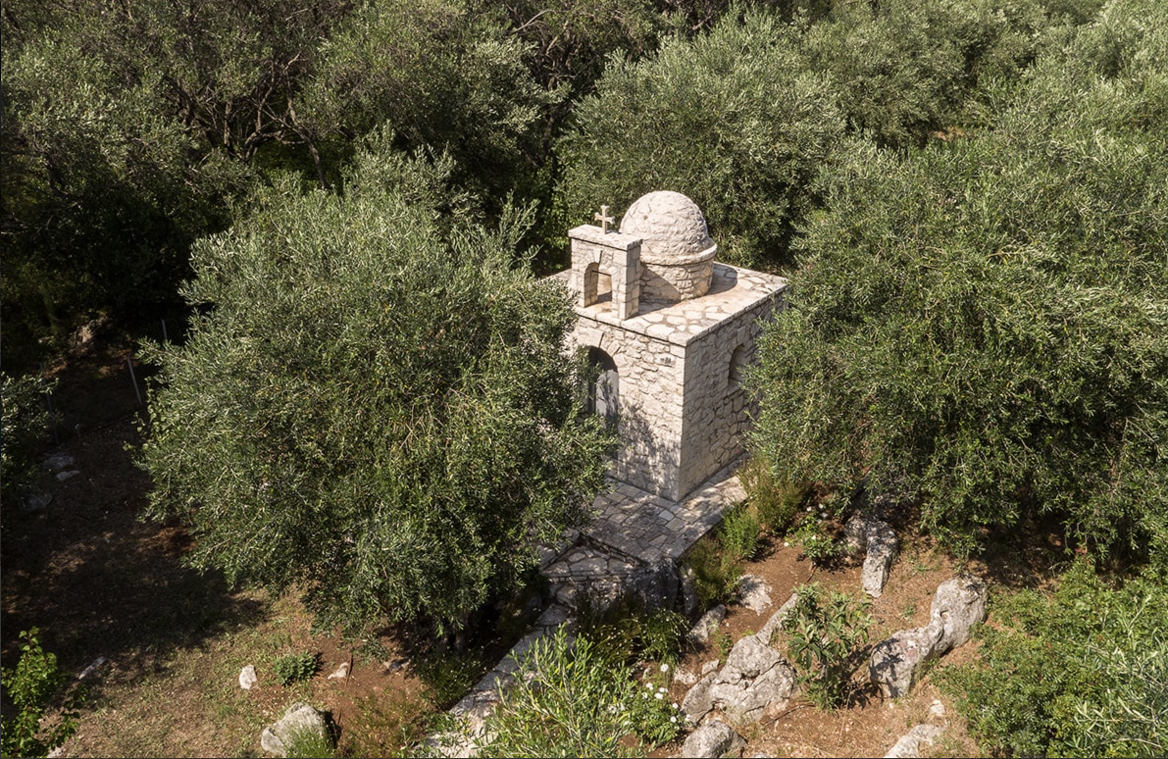 Stone chapel surrounded by dense green trees and bushes, with a stone pathway leading to it.