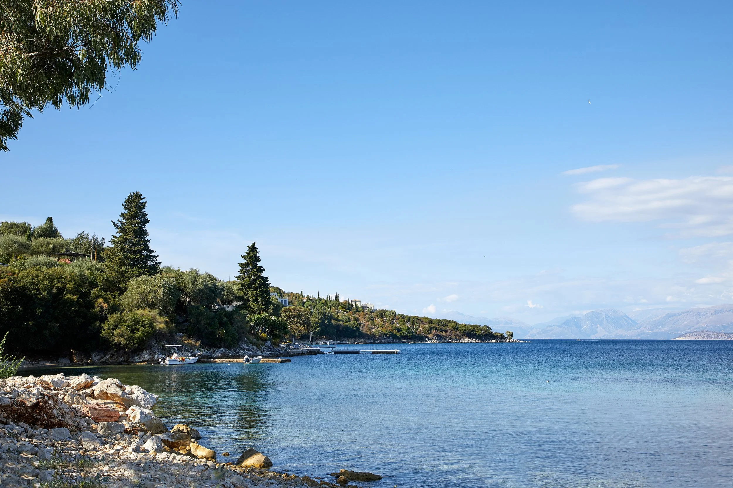 A peaceful lakeside scene with rocky shore, green trees, and calm water under a clear blue sky.