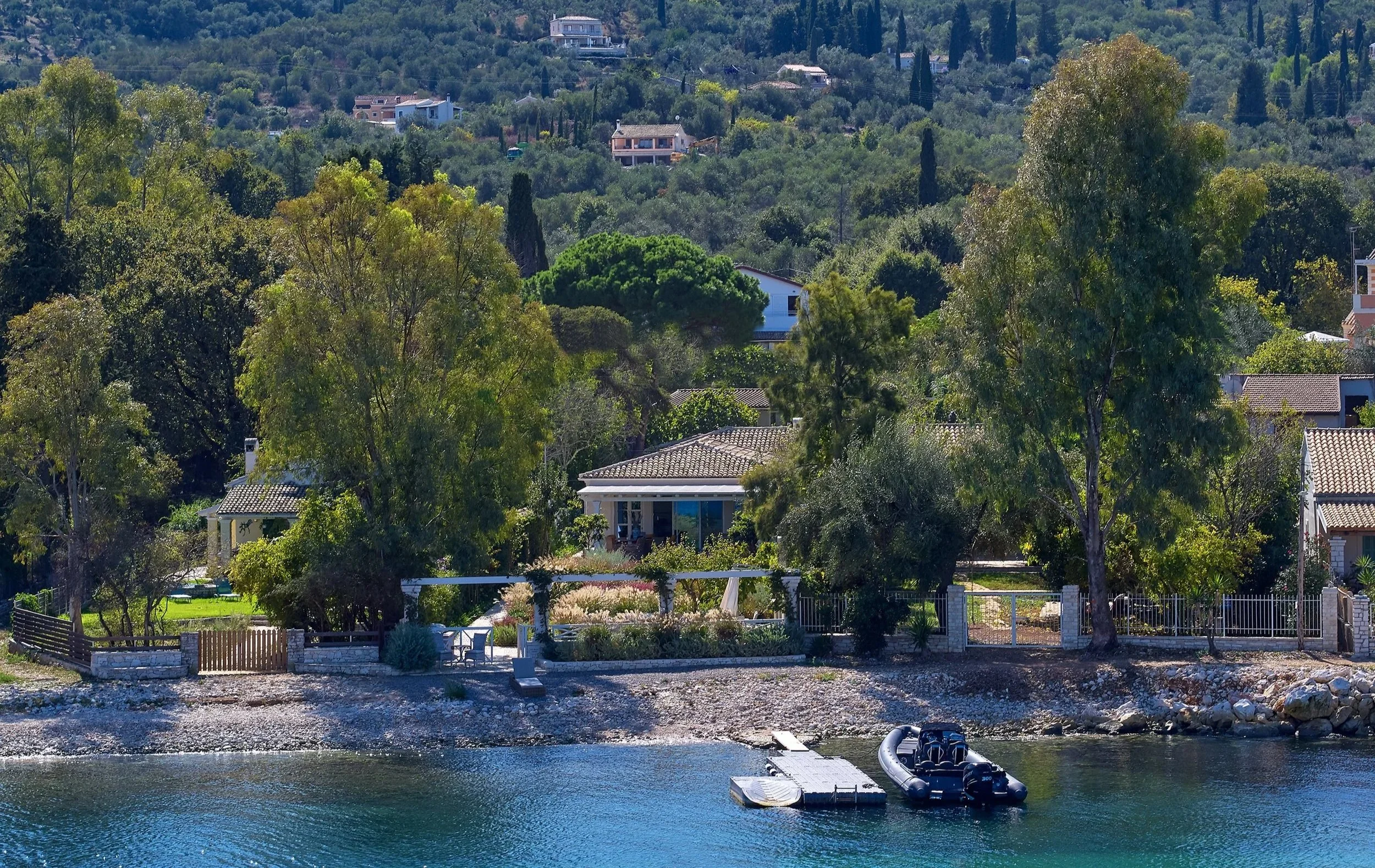 A lakeside view with houses surrounded by trees, a dock with a speedboat, and a hillside in the background.