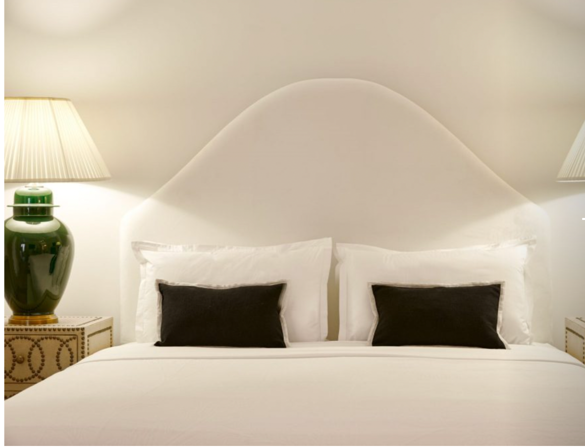 A neatly made bed with white sheets, two white pillows with black pillowcases, and a uniquely shaped white headboard. Two bedside tables with beige patterns are on either side, each holding a green ceramic lamp with beige shades.
