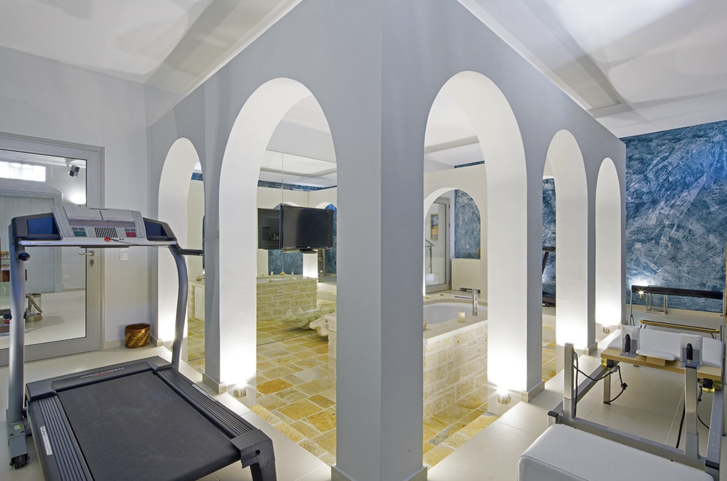 Modern wellness or spa room with white arches, exercise equipment, a small heated pool, and a textured blue wall.
