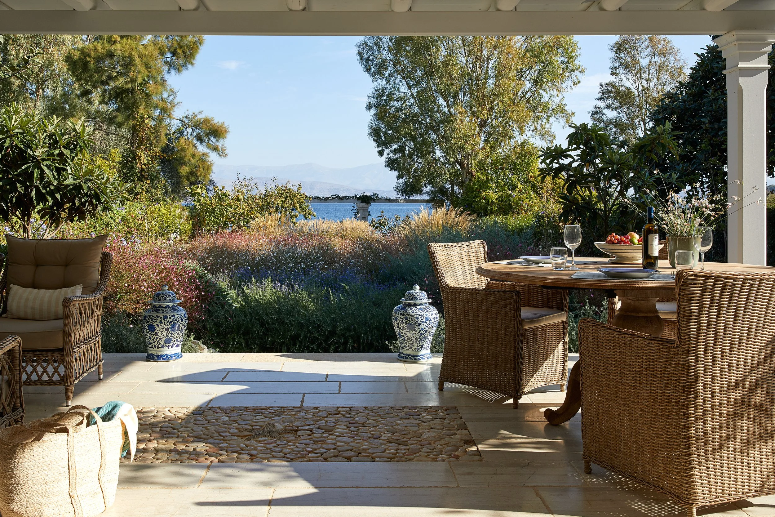 Outdoor patio with wicker chairs and a round table set with wine glasses, a bottle of wine, fruit, and flowers, overlooking a garden with colorful flowers, trees, and a view of water and mountains in the distance