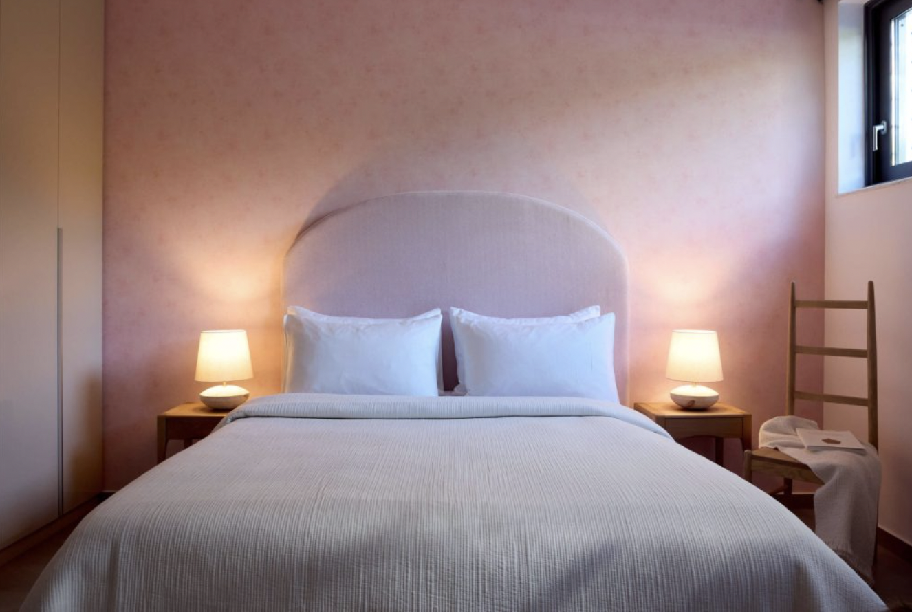 A cozy bedroom with a bed featuring a pink upholstered headboard, white bedding, and two white pillows. There are two matching bedside tables with lamps, and a wooden chair with a towel and some papers beside a window in the corner.