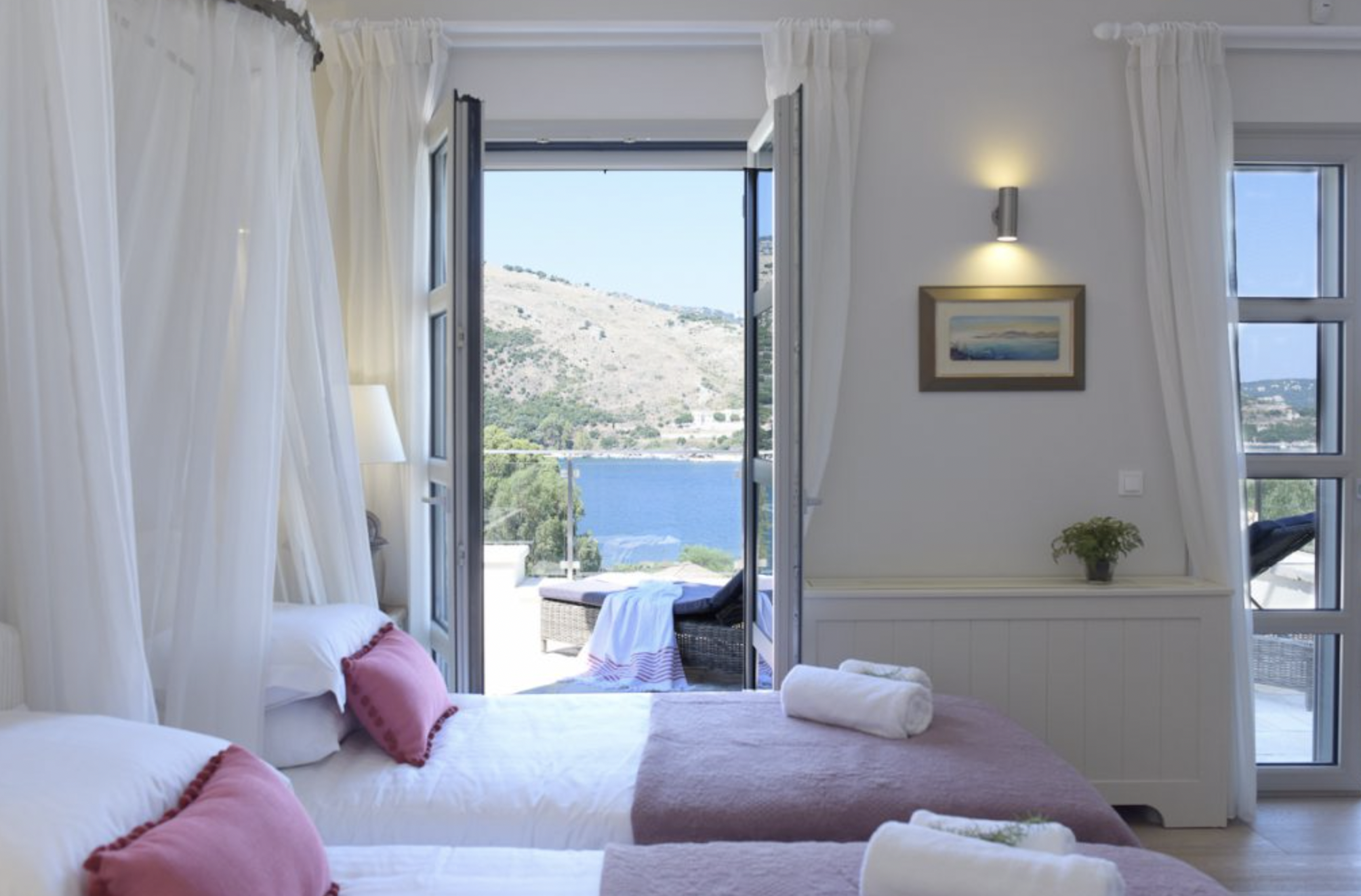 Bedroom with two beds, pink pillows, white linens, open balcony doors revealing a lake, a mountain, and clear sky, curtains, a framed painting, a wall-mounted light, a small potted plant.