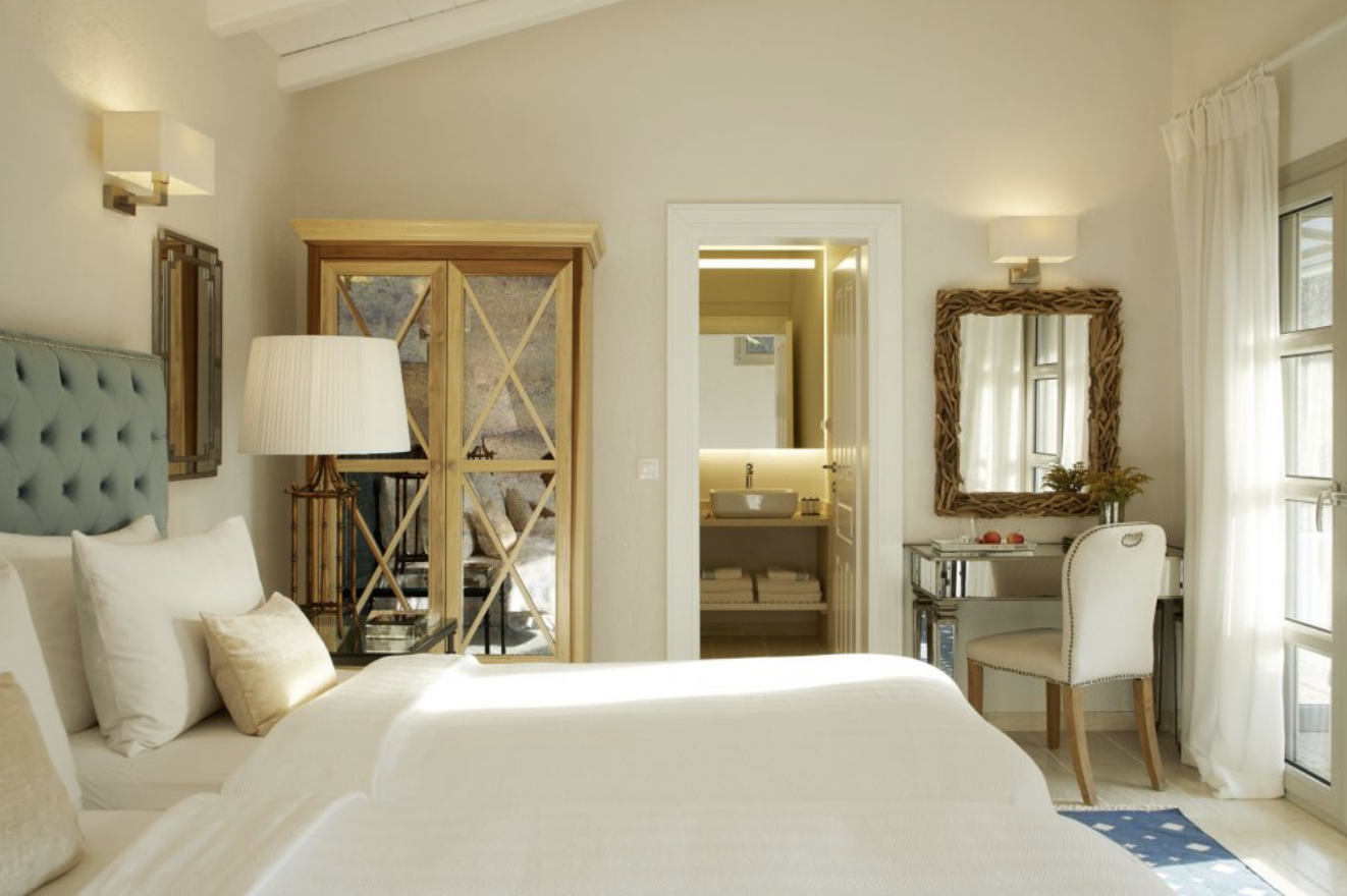 A cozy hotel room with two beds, a large headboard, a bedside table with a lamp, a wooden wardrobe, and a small desk with a chair, mirror, and window with white curtains.