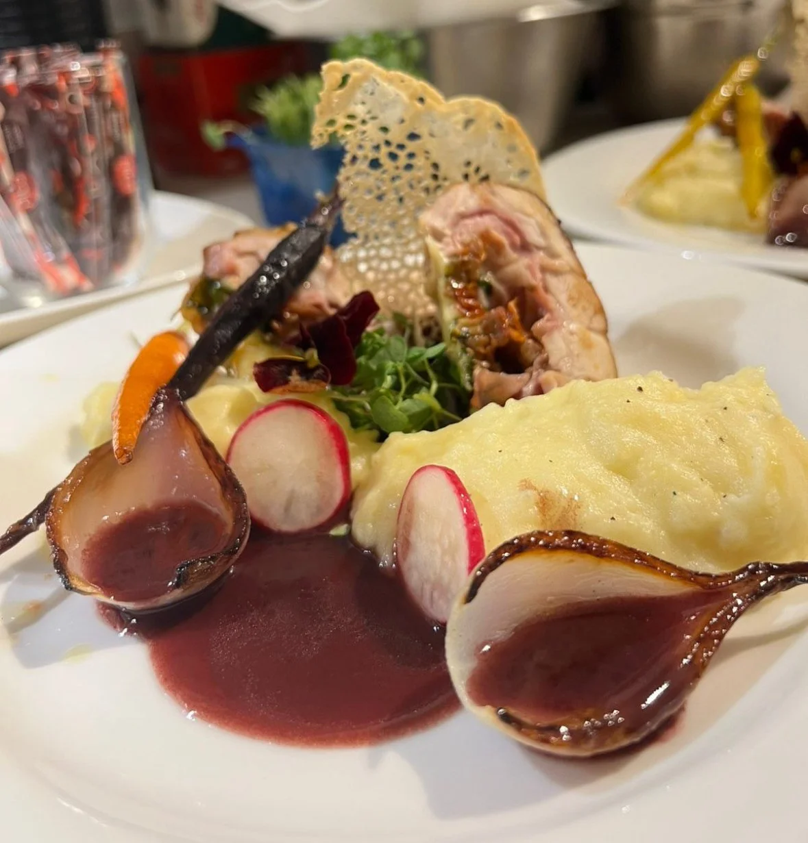 A plated gourmet meal featuring mashed potatoes, radish slices, meat with a dark sauce, roasted vegetables including carrots and garlic, a small green salad, and a decorative cheese crisp.