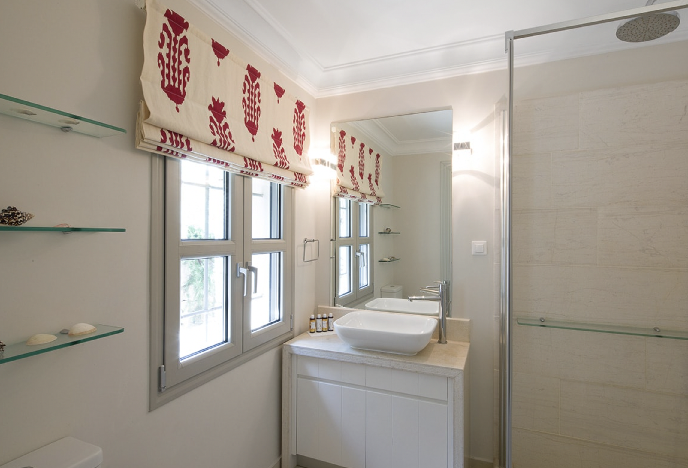 A bathroom with two windows, a mirror, a sink with three small bottles, shell decorations on glass shelves, and a walk-in shower with a rainfall showerhead.